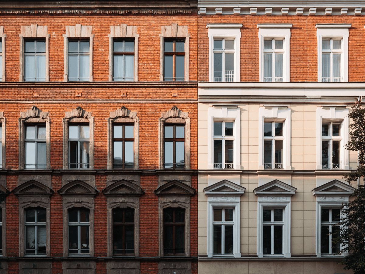 Facade of two adjoining buildings, one with red bricks and dark windows, the other with light plaster and white-framed windows, under soft daylight.