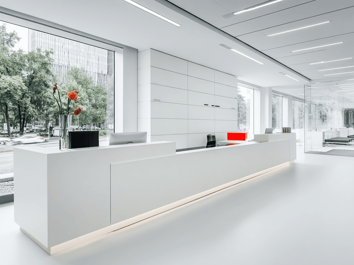 Modern, minimalist office reception with a sleek white desk, flower vase, and large windows offering a view of trees and city buildings.