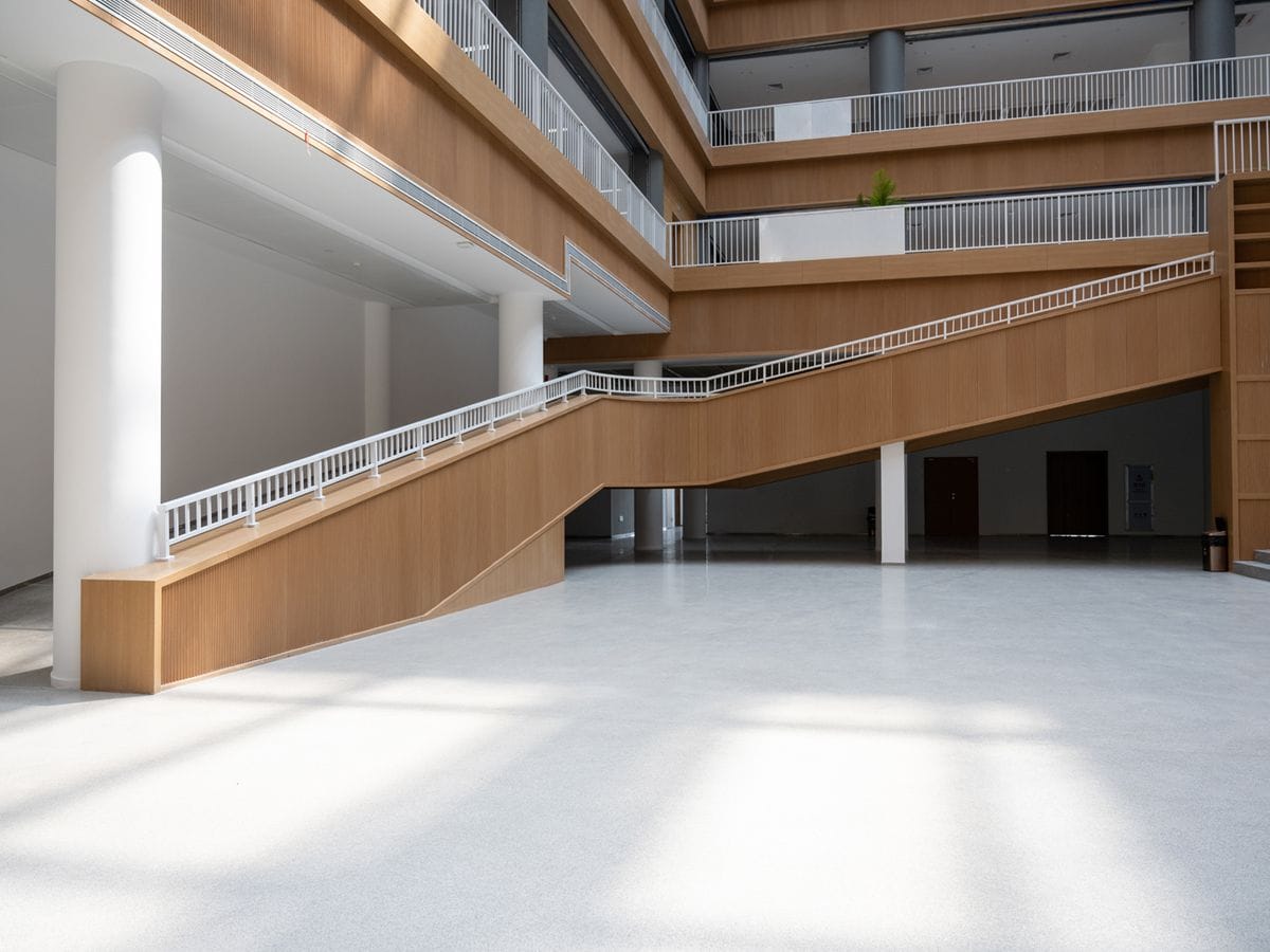 Spacious modern atrium with wooden accents, white railings, and a wide staircase, illuminated by natural light from large windows.