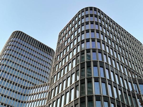 Two modern, curved glass and steel buildings under a clear blue sky, with a perspective from below emphasizing their height and sleek design.