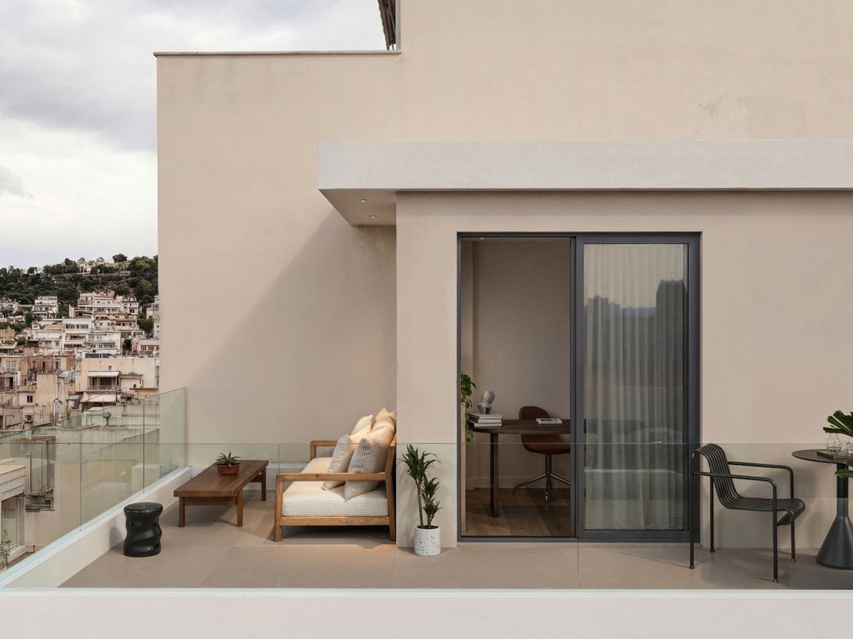Modern rooftop patio with glass railings, beige walls, a cozy sofa, potted plants, and a sliding door leading to an interior office space.