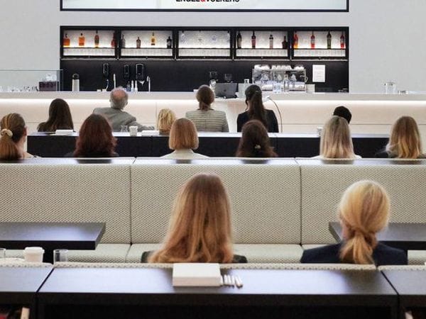Audience seated in rows facing a bar area in a bright, modern space. People are listening to a presentation.
