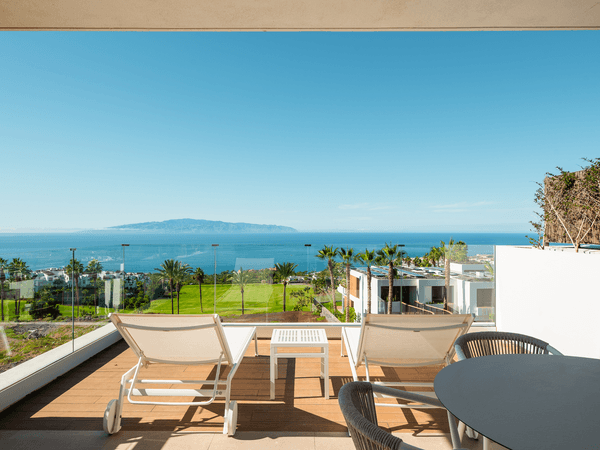 Terrace with lounge chairs and a table overlooking a green lawn, palm trees, and ocean with an island in the distance under a clear blue sky.