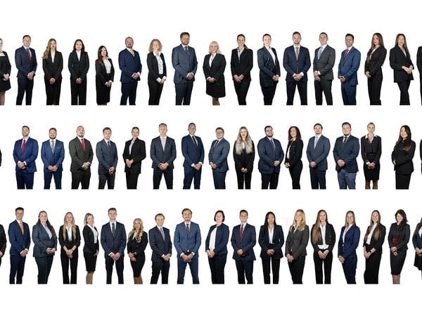Group photo of the entire Engel & Völkers team. Dozens of men and women in business suits stand in several horizontal rows against a white background, with the Engel & Völkers logo prominently displayed in the center.