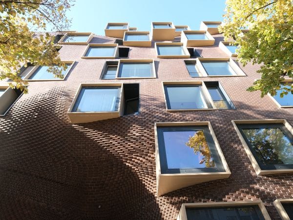 Low-angle view of a brick building with staggered box-like windows and autumn trees framing the sunlit facade.