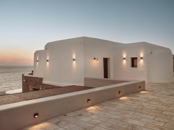 Εξωτερική άποψη μιας λευκής μεσογειακής βίλας με πέτρινη βεράντα που βλέπει στον ωκεανό στο ηλιοβασίλεμα.