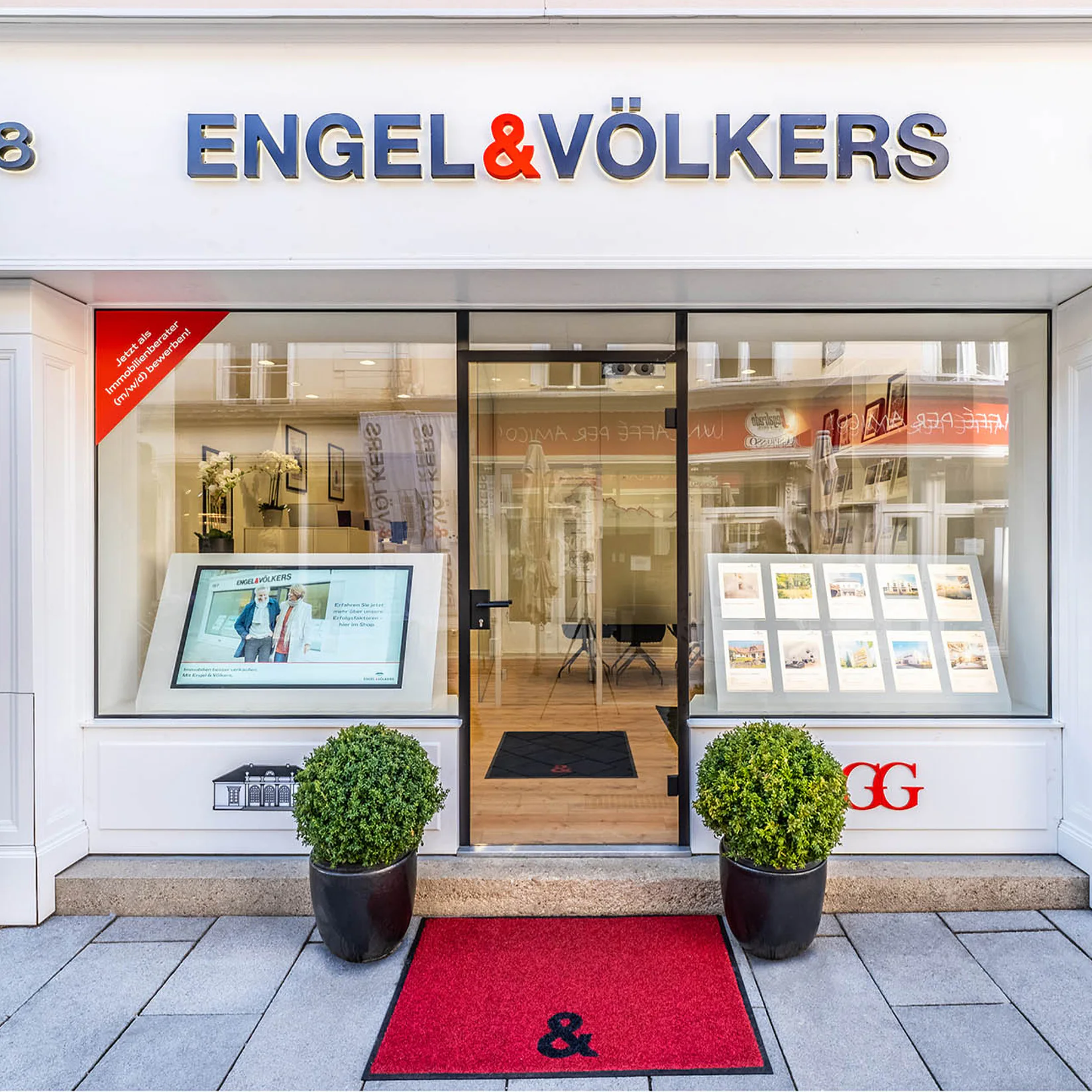 Engel & Völkers real estate storefront with a glass facade, white trim, and the company's blue logo above the entrance. Two potted plants flank a red welcome mat.