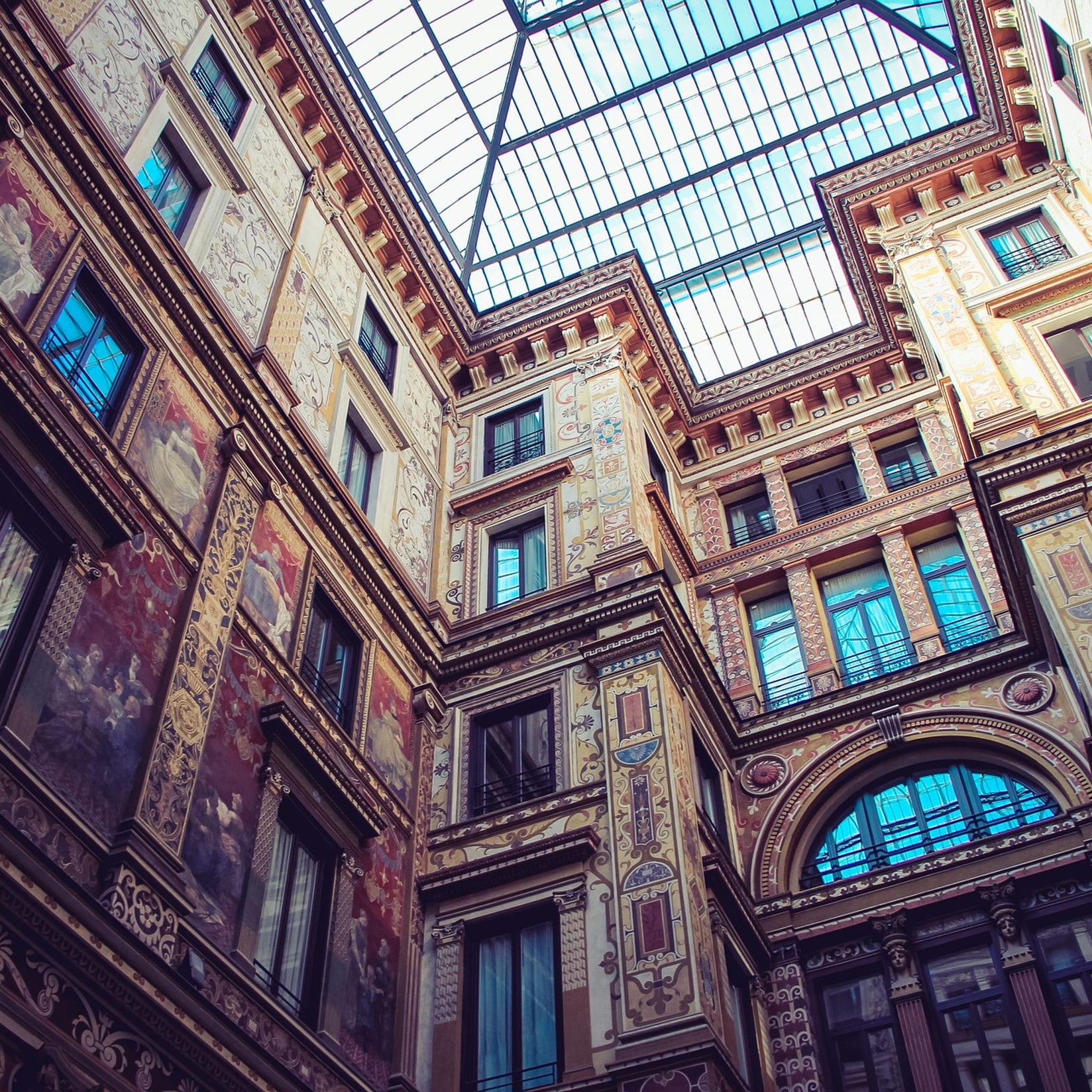 Verzierter Gebäudeinnenraum mit einer Glasdecke. Die Wände sind mit Wandmalereien und dekorativen Mustern in Rot-, Beige- und Brauntönen versehen. Die Fenster spiegeln den Himmel wider.