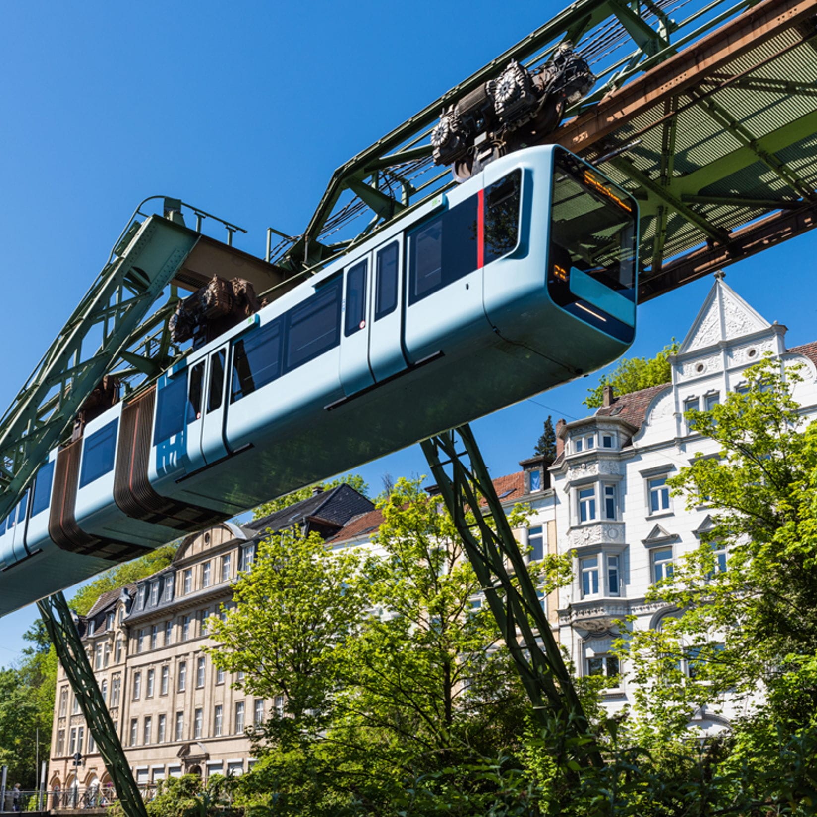 Ein hellblauer Schwebebahnwagen der Wuppertaler Schwebebahn fährt an einem sonnigen Tag über grüne Bäume und Gebäude.