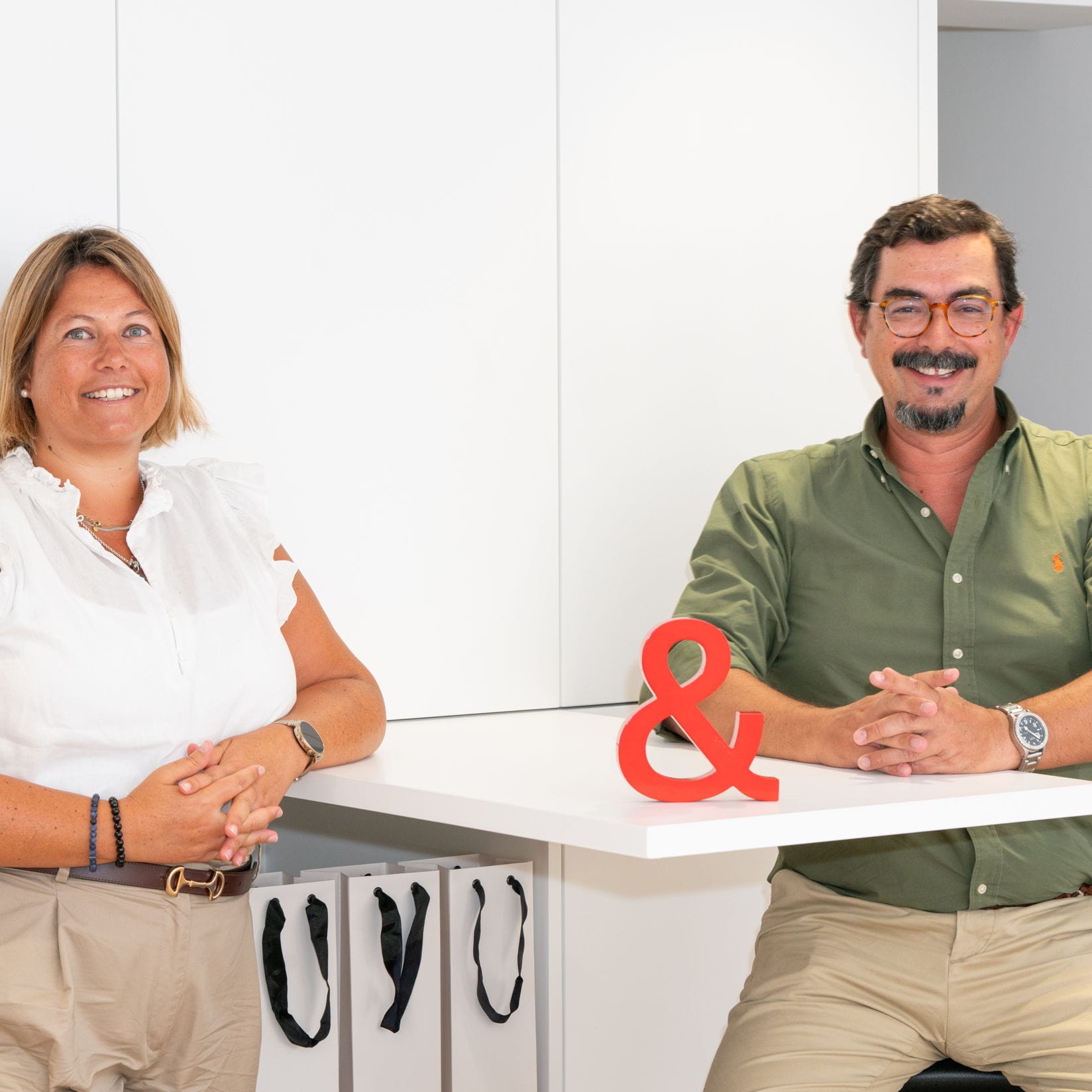 Real estate agents, a woman in white and a man in green, lean on a white table with a red ampersand.