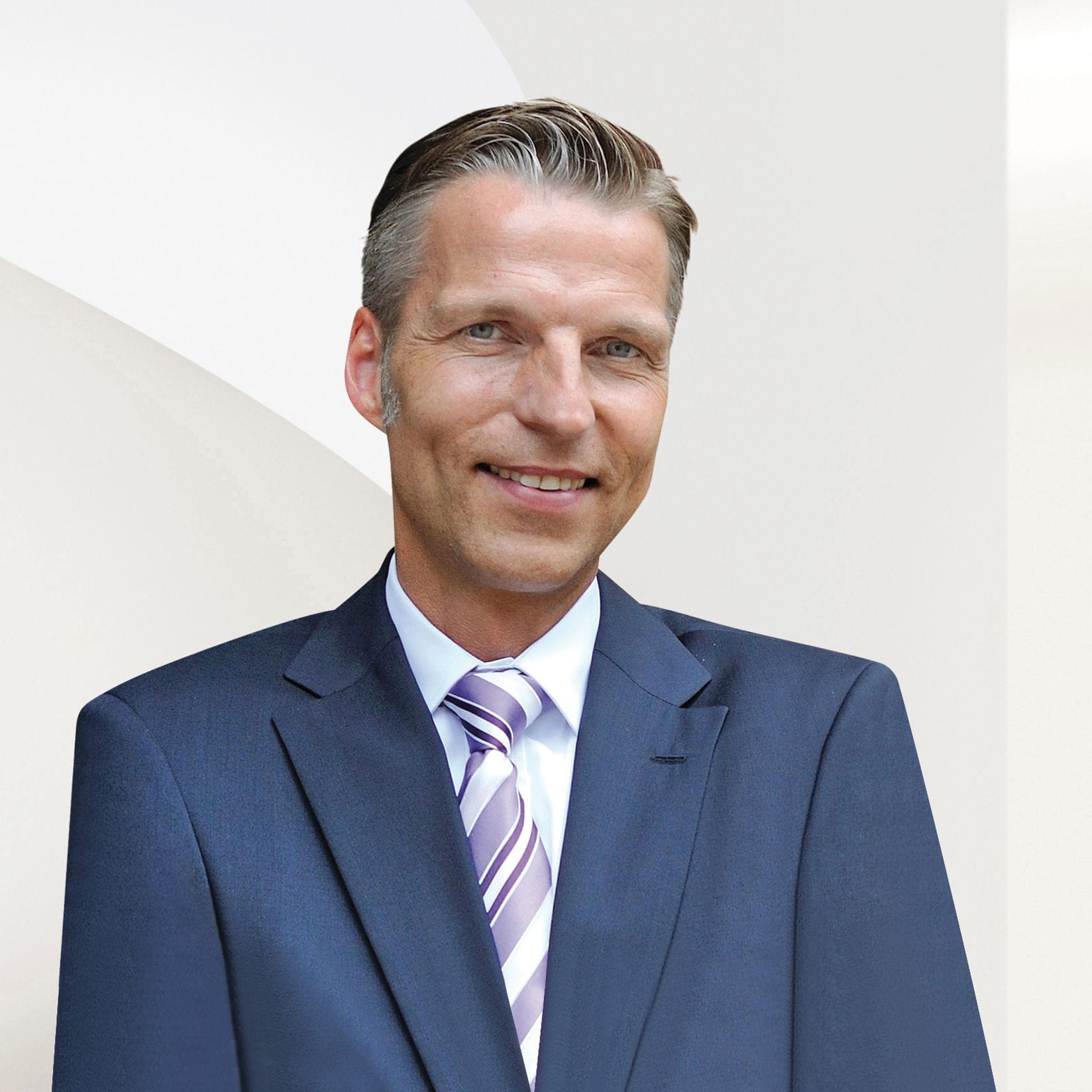 Ein lächelnder Mann in einem blauen Anzug und einer gestreiften Krawatte steht vor einem weißen Hintergrund.