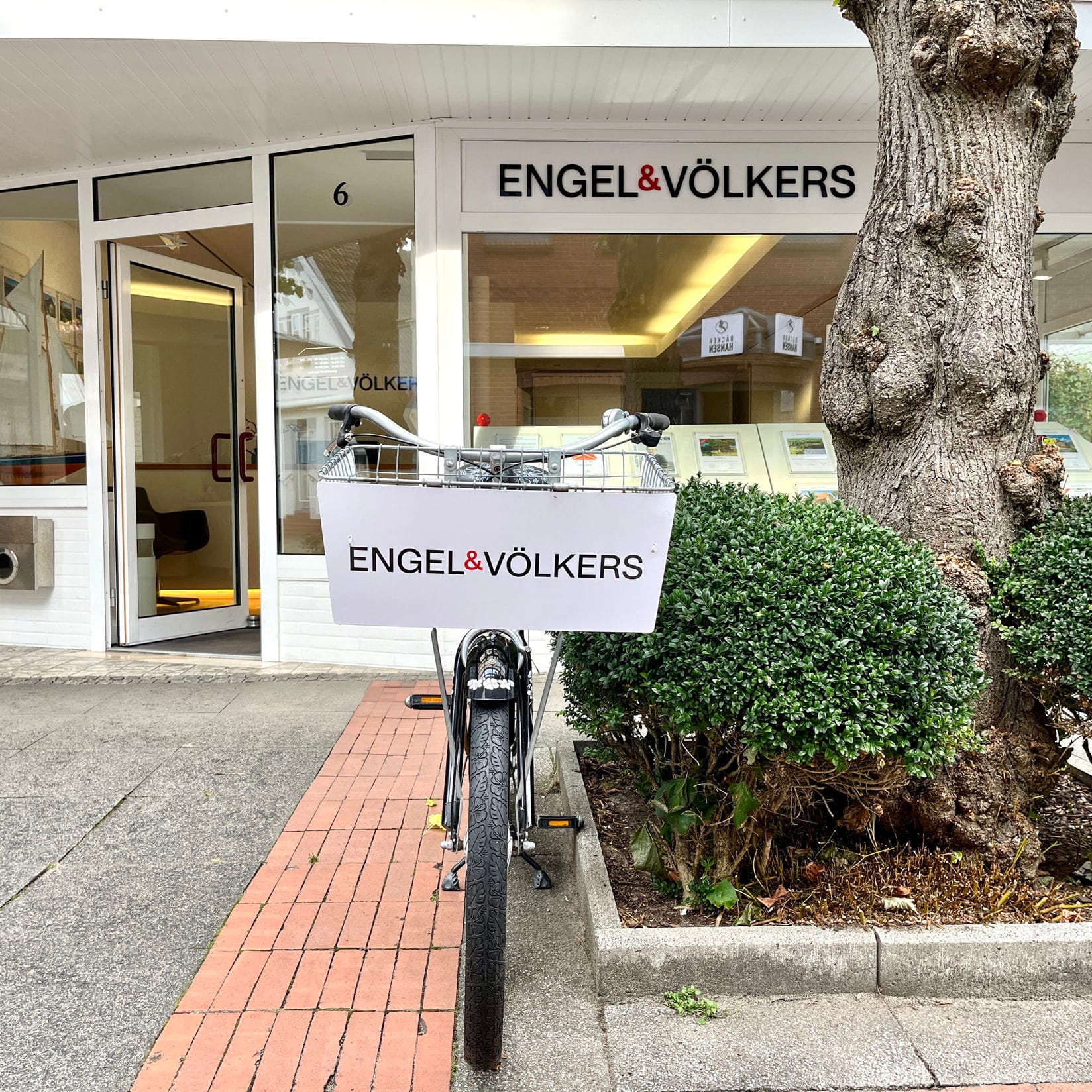 Ein Fahrrad mit einem Engel & Völkers Schild im Korb steht vor deren Büro. Das Gebäude hat große Fenster und einen weißen Rahmen.