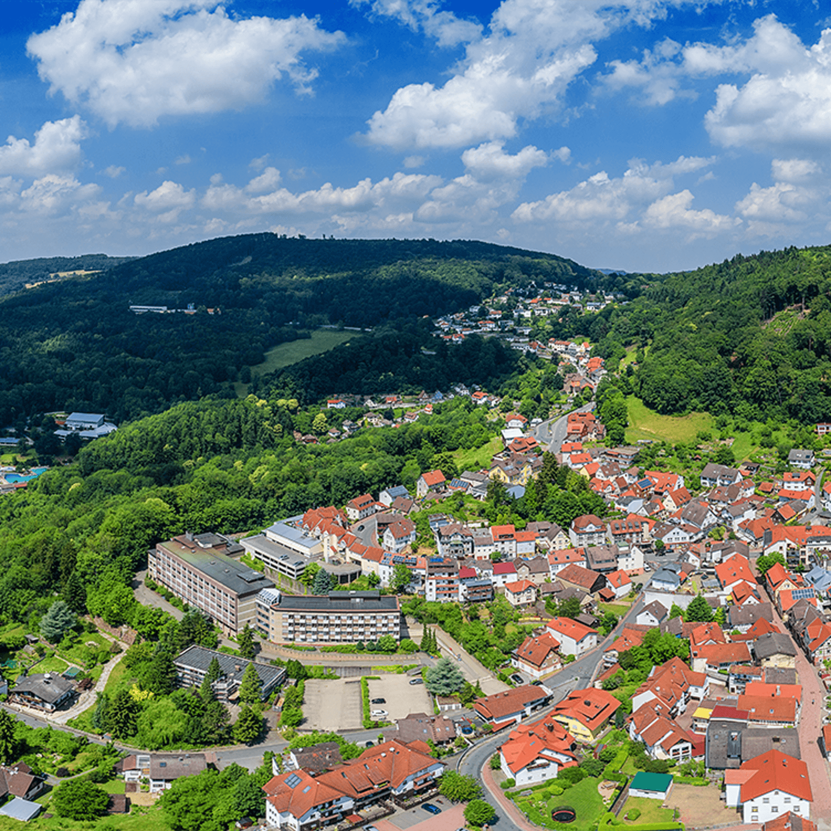Luftaufnahme einer Stadt mit rot gedeckten Gebäuden, eingebettet in grüne Hügel unter einem blauen Himmel mit weißen Wolken.