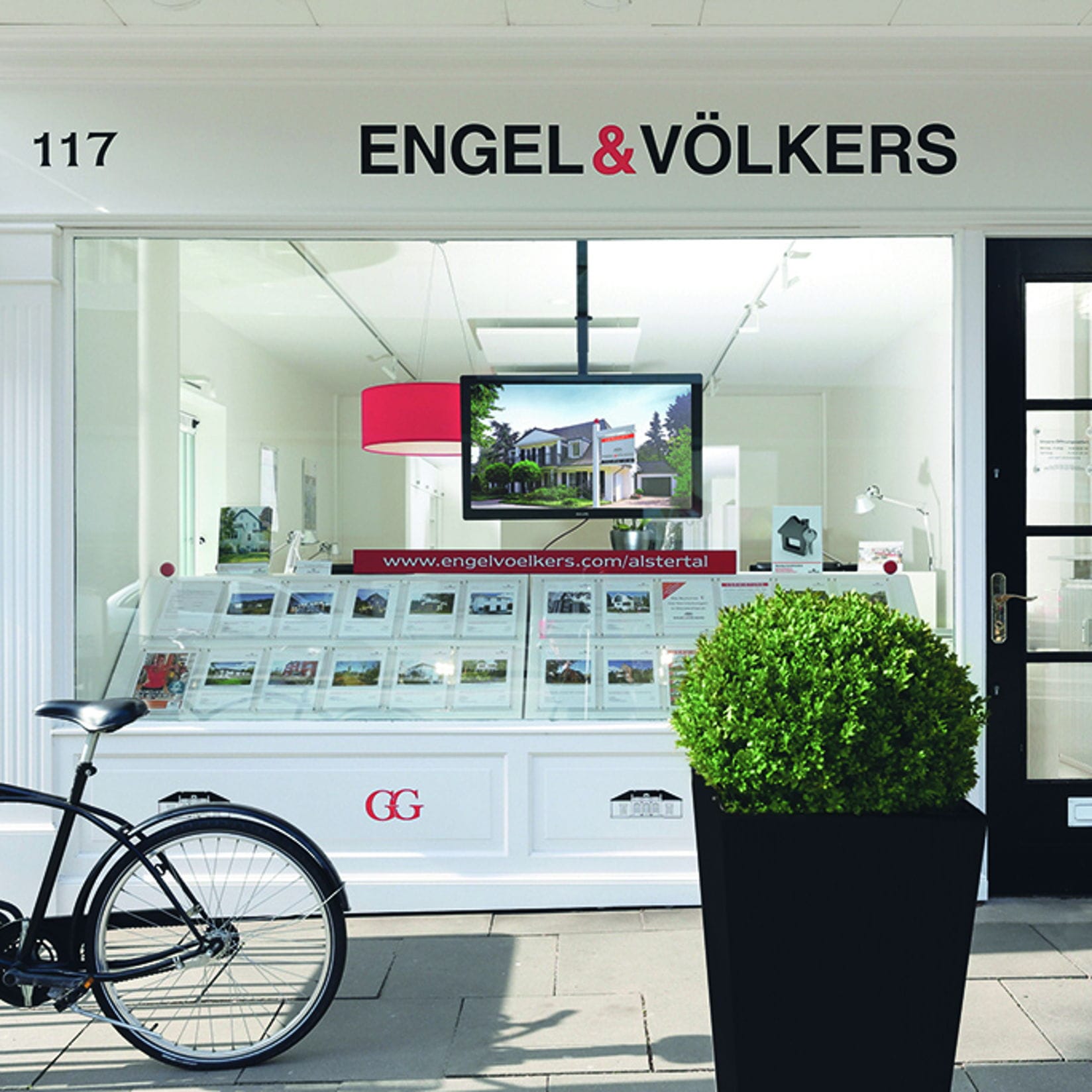 Engel & Völkers real estate storefront with a black bicycle parked outside. The window displays property listings and a TV screen.