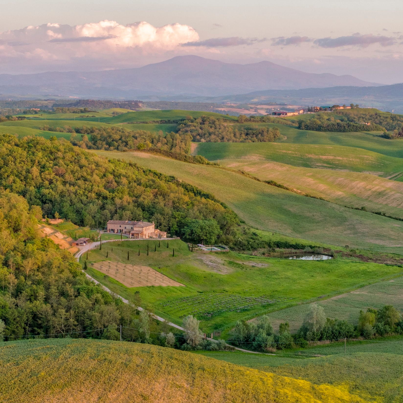 Vista panoramica di una villa toscana circondata da verdi colline ondulate e montagne lontane sotto un cielo color pastello.