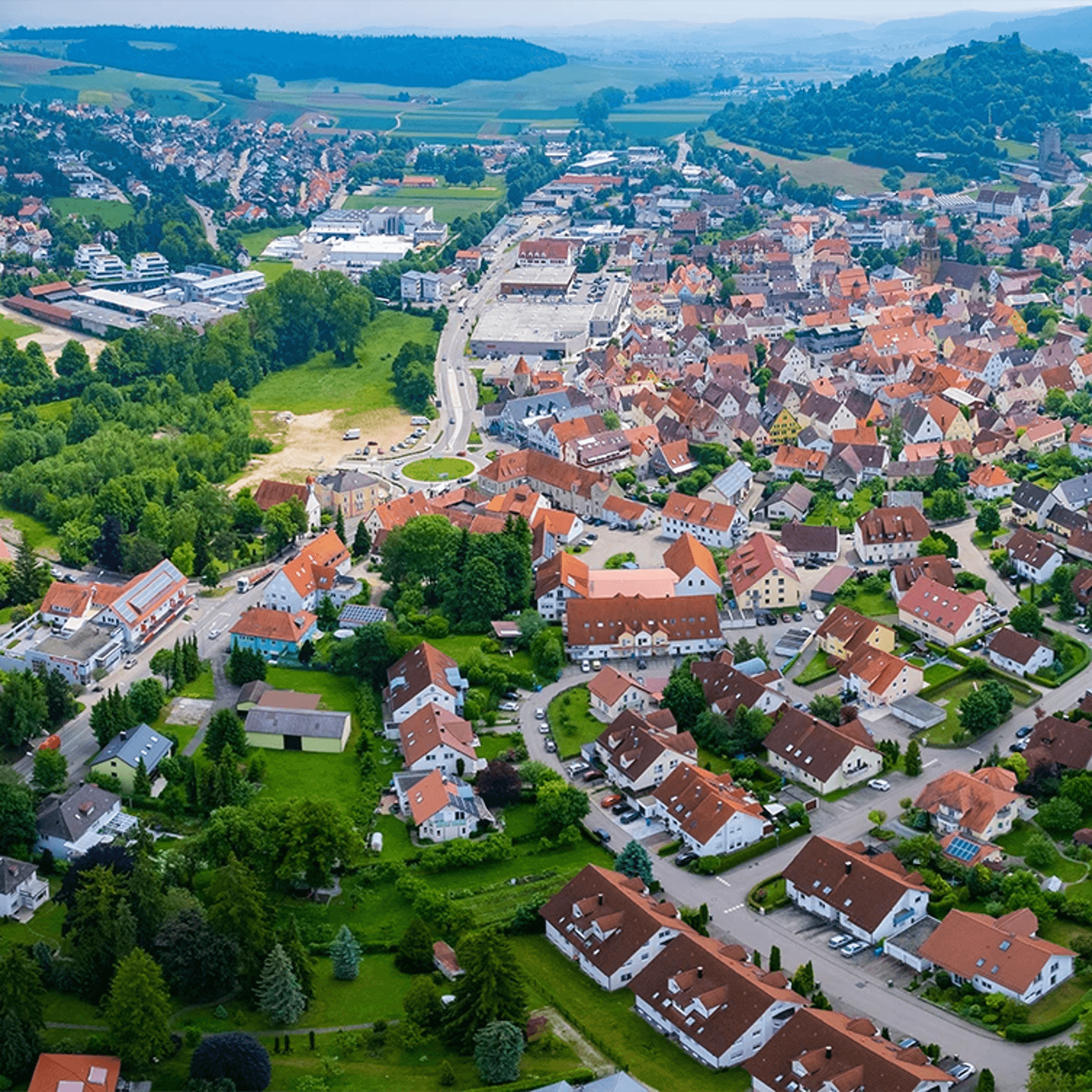 Aerial view of a European town with red-roofed houses, green trees, and rolling hills in the background.