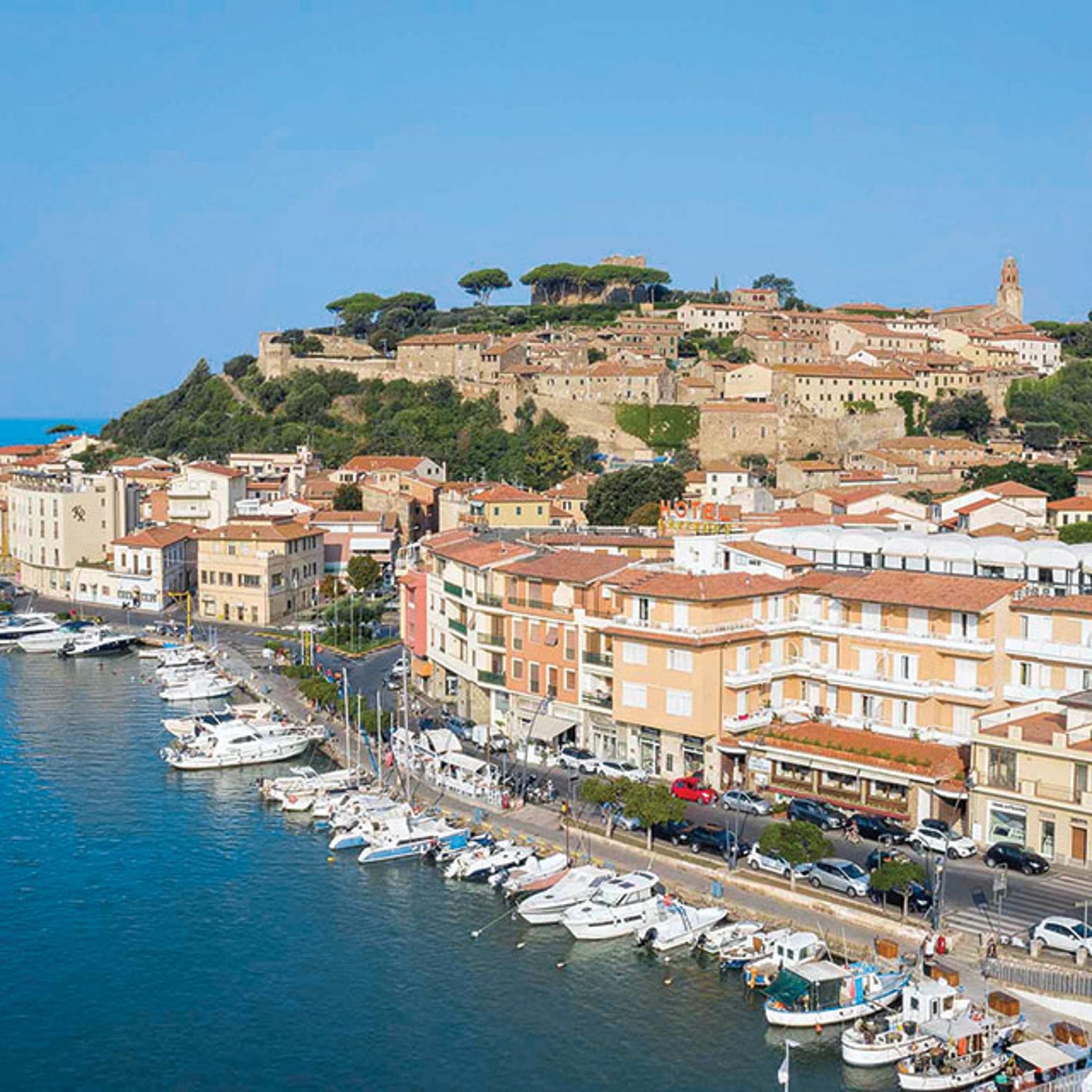 Coastal town with boats docked along the waterfront, buildings with red tile roofs, and a hilltop fortress under a blue sky.