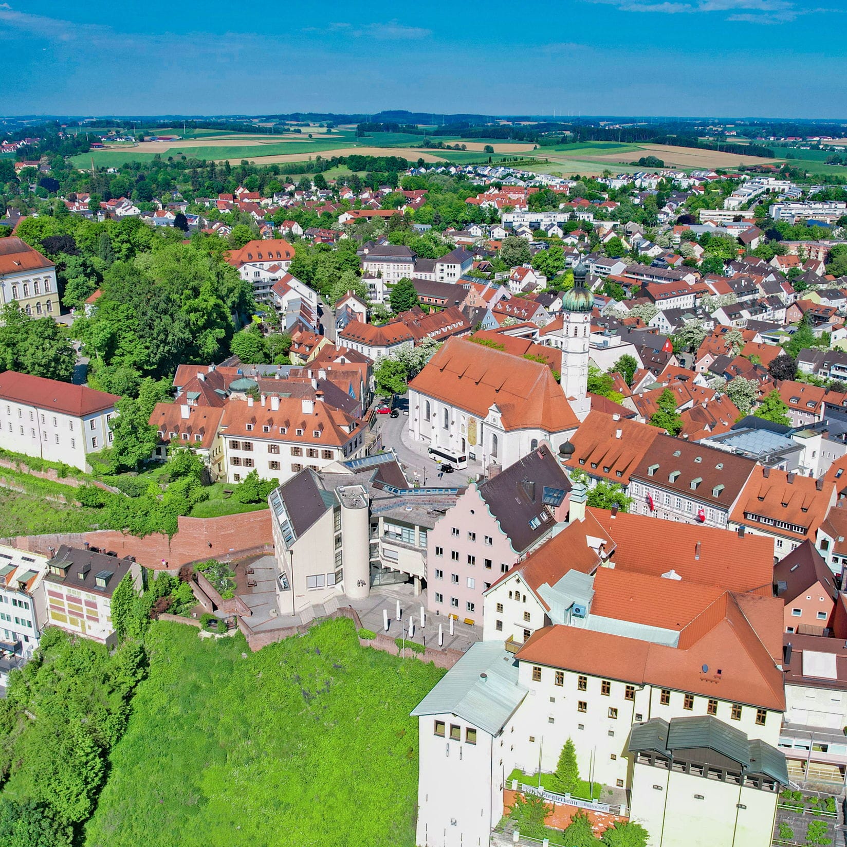 Luftaufnahme einer europäischen Stadt mit roten Ziegeldächern, grünen Bäumen und blauem Himmel.