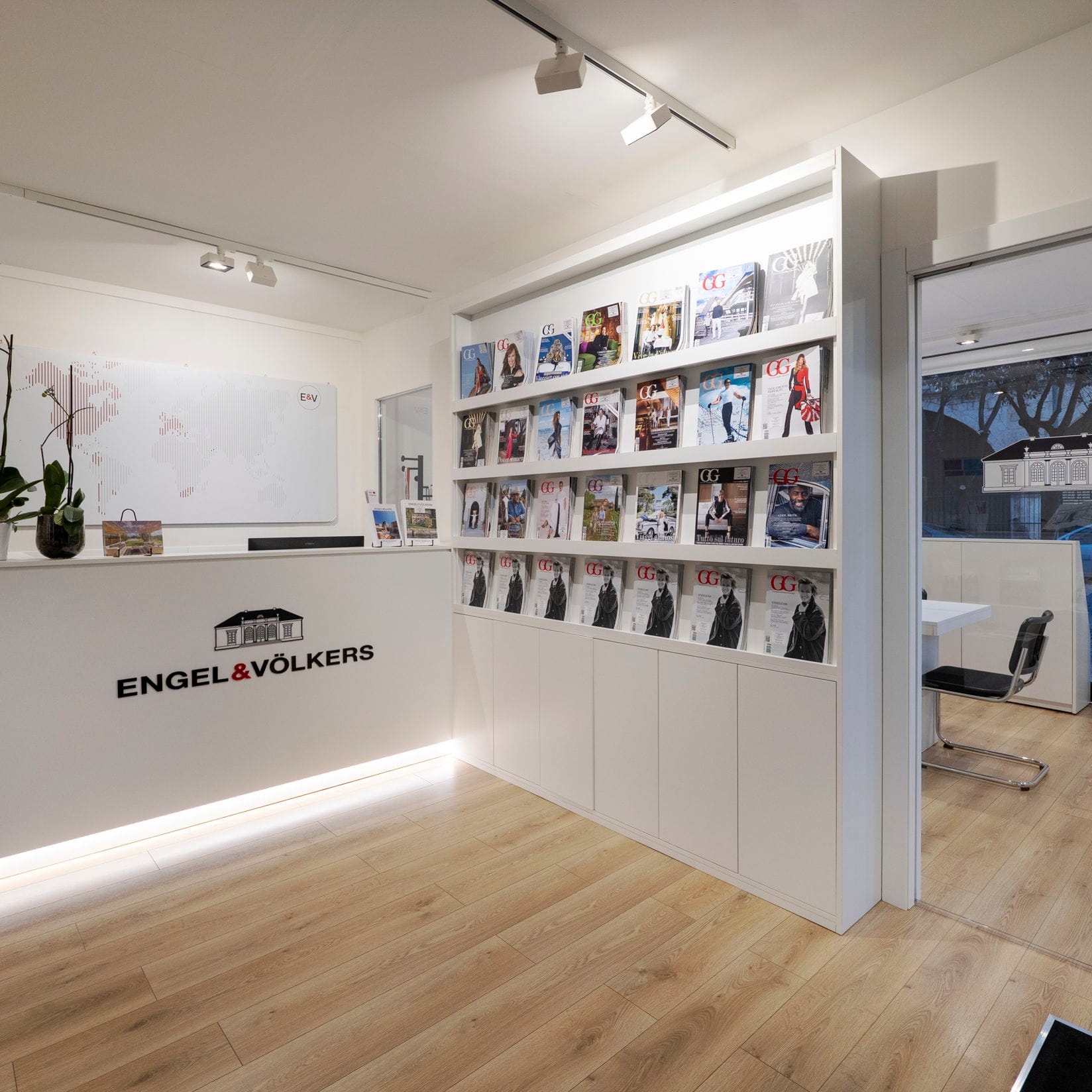 Interni dell'ufficio Engel & Völkers con una reception bianca, orchidea e portariviste. Una porta conduce a una sala riunioni.