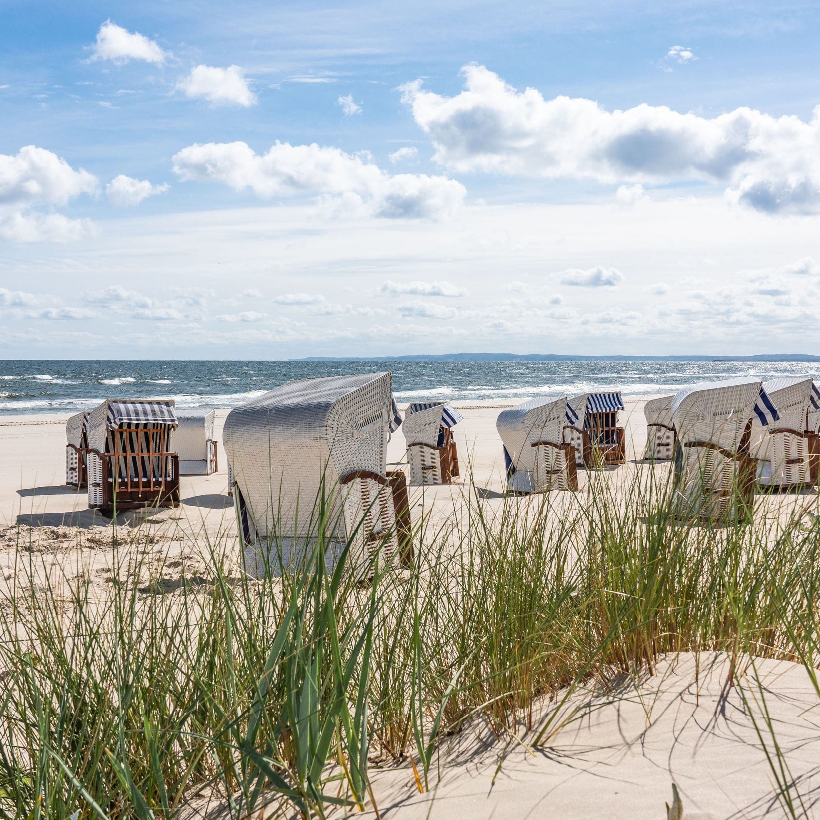 Beach scene with rows of white wicker beach chairs on sand, ocean waves, and blue sky with white clouds.