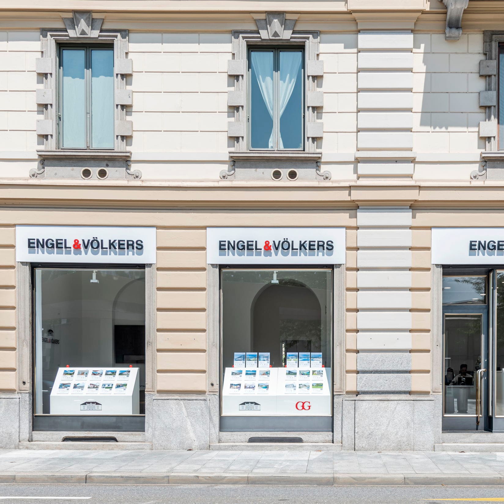 Engel & Völkers real estate office with three large windows displaying property listings on a sunny day.