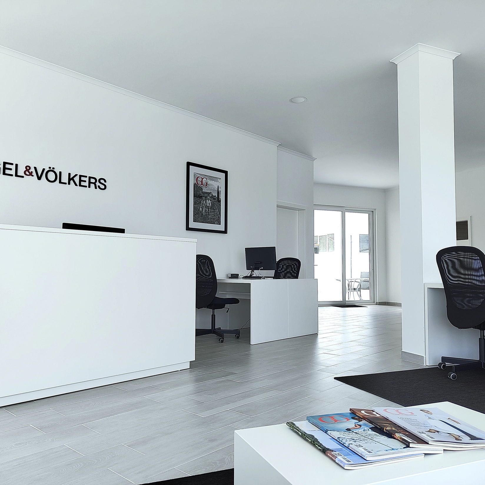 Engel & Völkers Büro-Interieur mit weißem Empfangstresen, schwarzem Logo und Zeitschriften auf einem weißen Tisch.