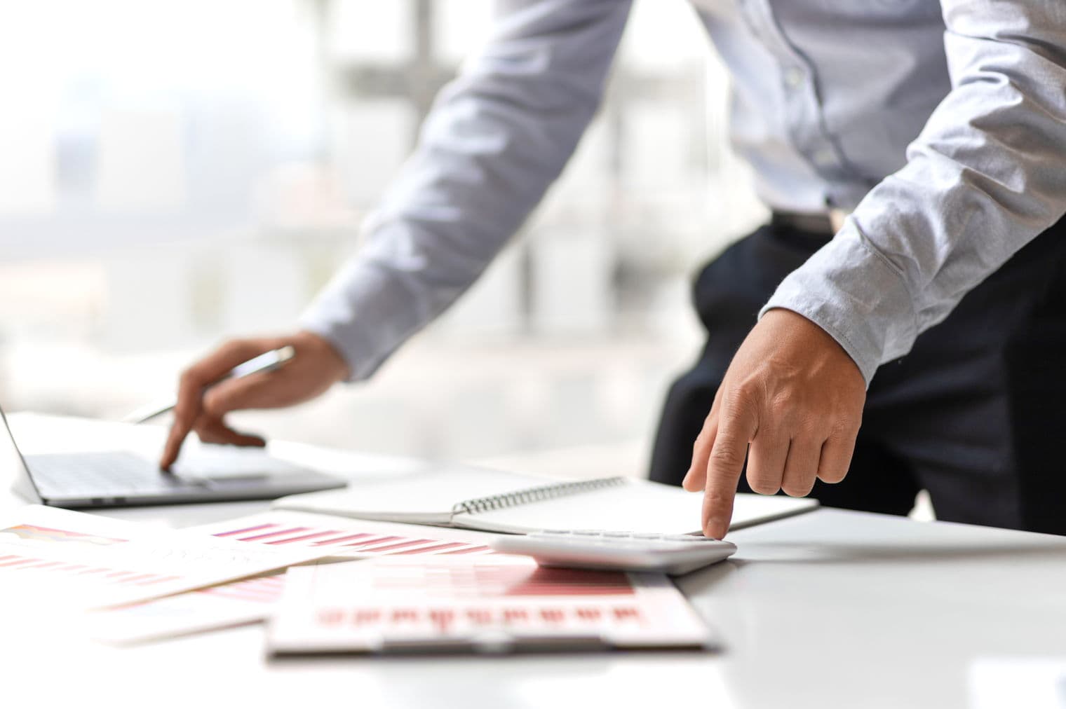 Eine Person in einem hellblauen Hemd benutzt einen weißen Rechner auf einem weißen Schreibtisch mit Diagrammen und einem Laptop.