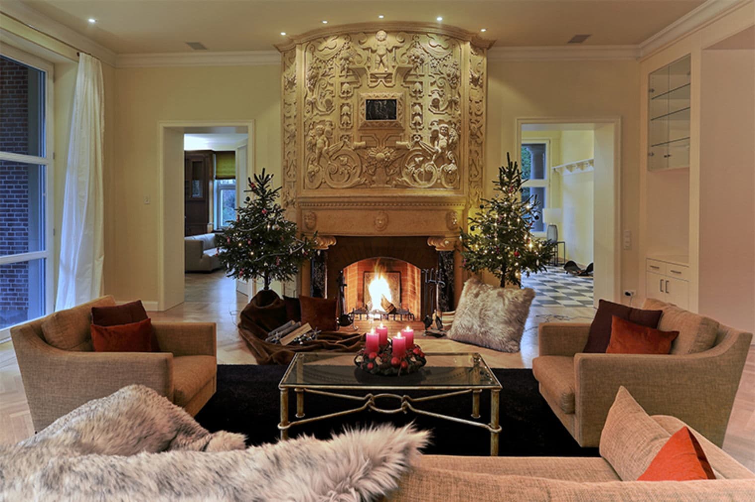 Living room with a large, ornate fireplace, two Christmas trees, and tan sofas. A glass coffee table sits on a black rug.