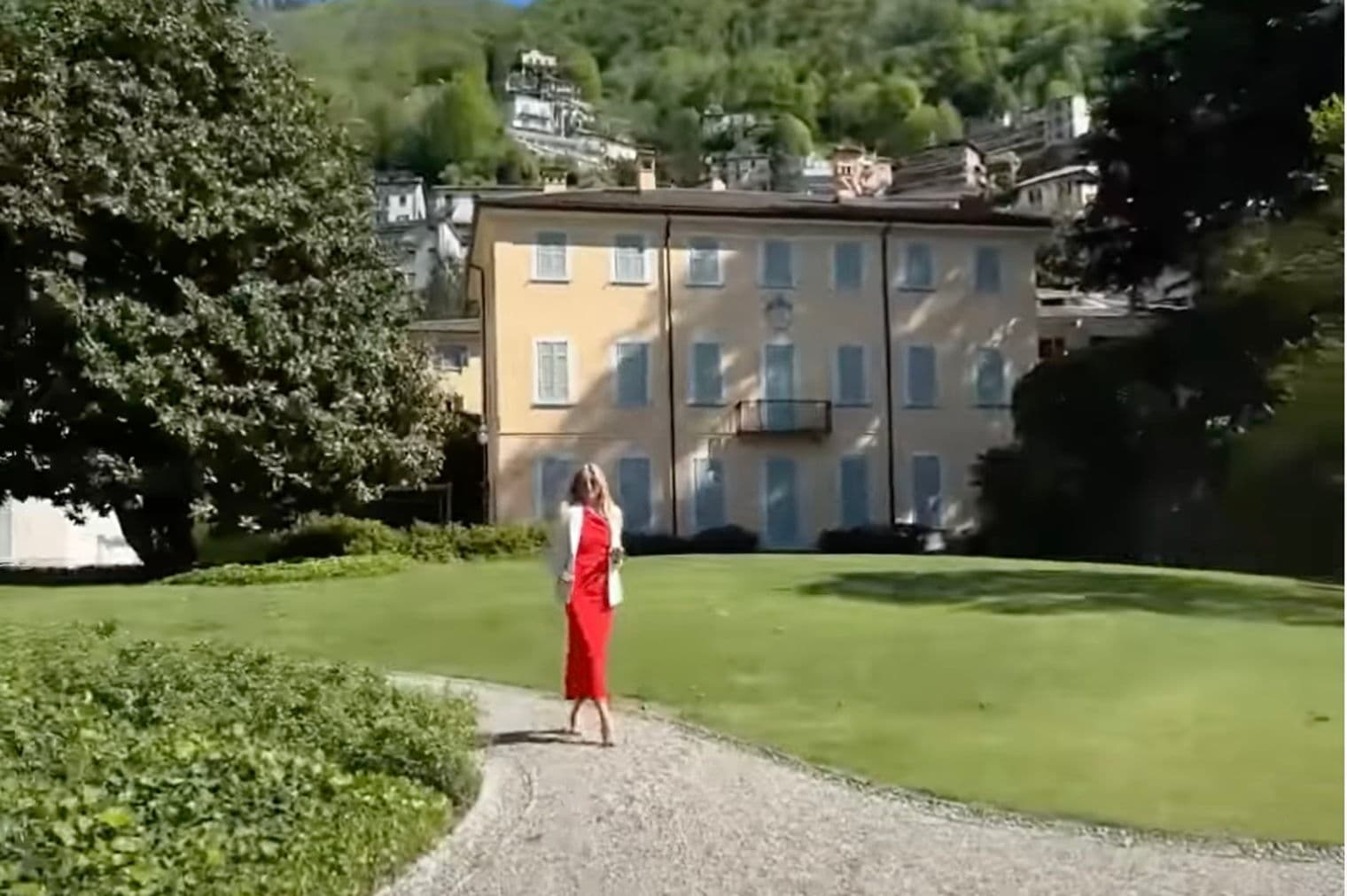 A woman in a red dress walks on a gravel path towards a large, yellow building with blue shutters, set against a green hillside.