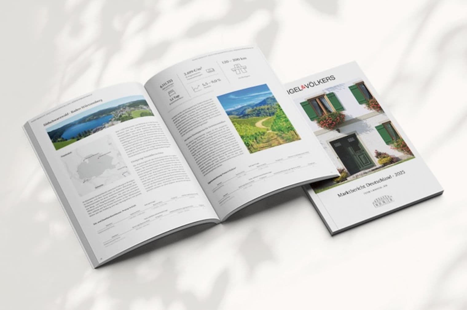 Geöffnete Immobilienbroschüre mit Landschaftsfotos, neben einer geschlossenen Broschüre mit einer Hausfassade auf einer weißen Oberfläche.