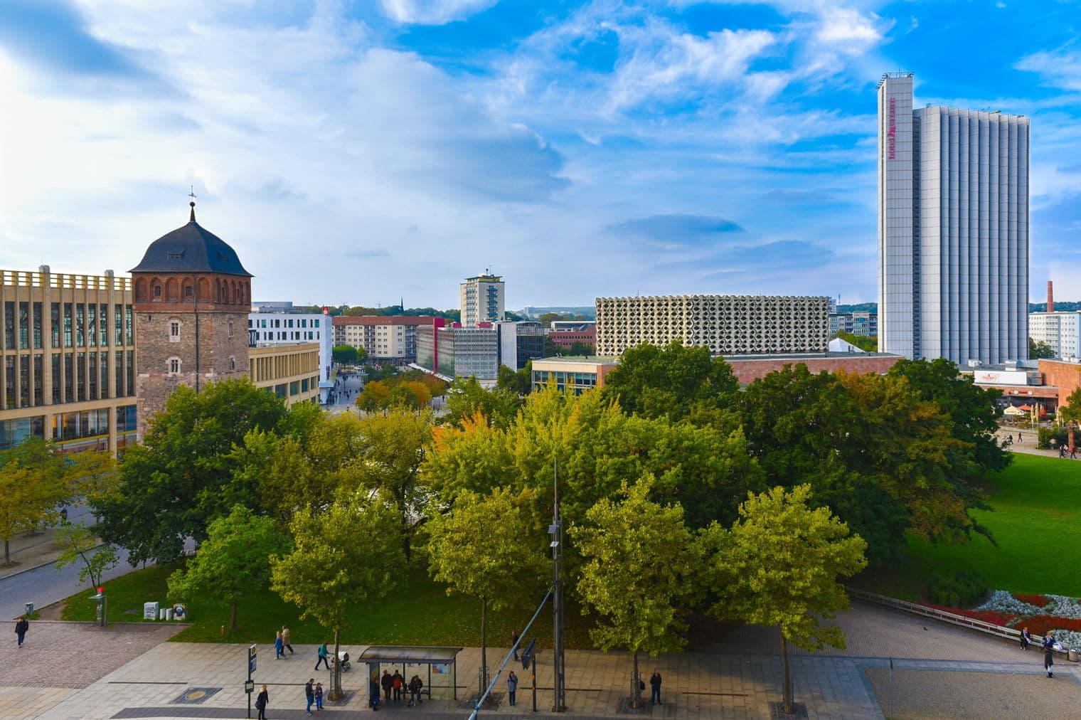 Stadtansicht von Magdeburg, Deutschland, mit einem historischen Turm, modernen Gebäuden, grünen Bäumen und einem blauen Himmel mit weißen Wolken.