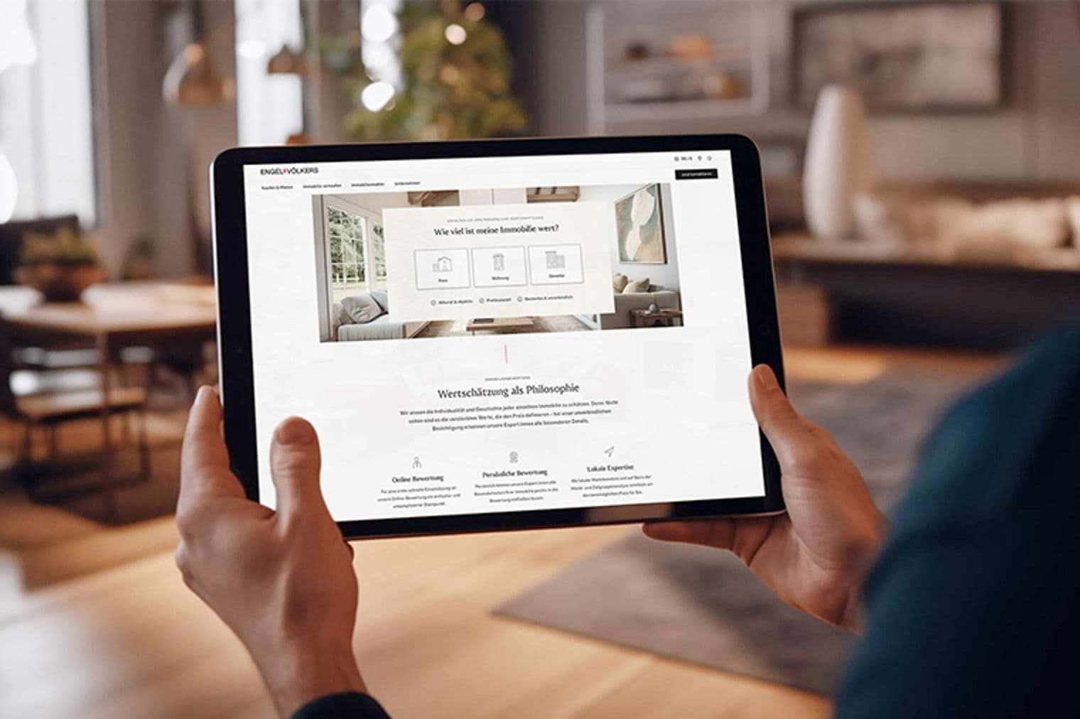 Hands holding a tablet displaying the Engel & Völkers real estate website in a blurred living room setting.