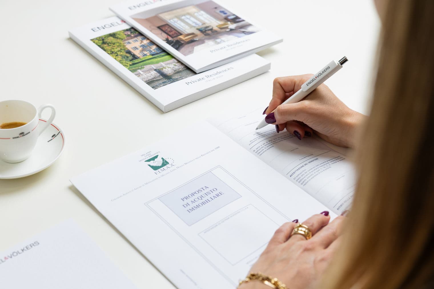 Der Makler unterzeichnet einen Immobilienkaufvorschlag mit einem Engel & Völkers Stift. Kataloge und eine Kaffeetasse stehen auf dem weißen Tisch.
