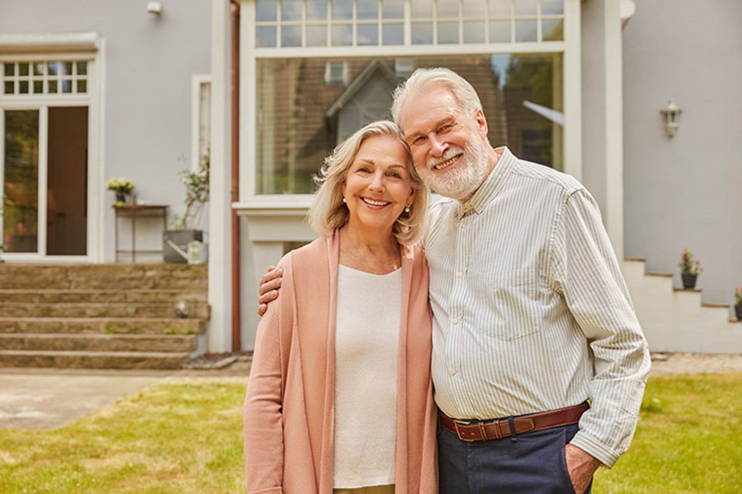 Smiling senior couple stands in front of a grey house with white trim and green lawn. The woman wears a pink cardigan.