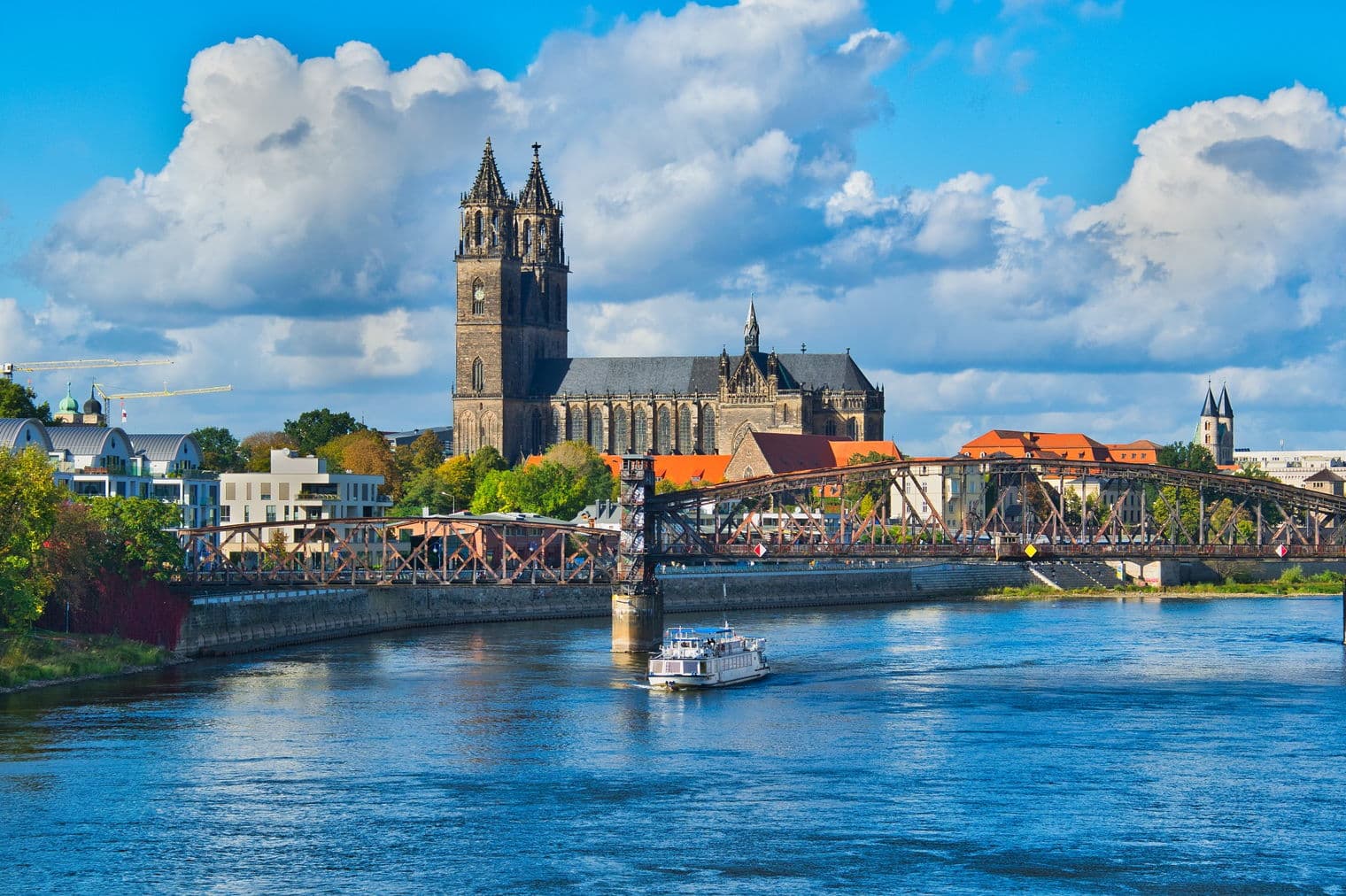 Blick auf den Magdeburger Dom und die Brücke über die Elbe, Deutschland, unter blauem Himmel mit weißen Wolken. Ein weißes Boot segelt auf dem blauen Wasser.