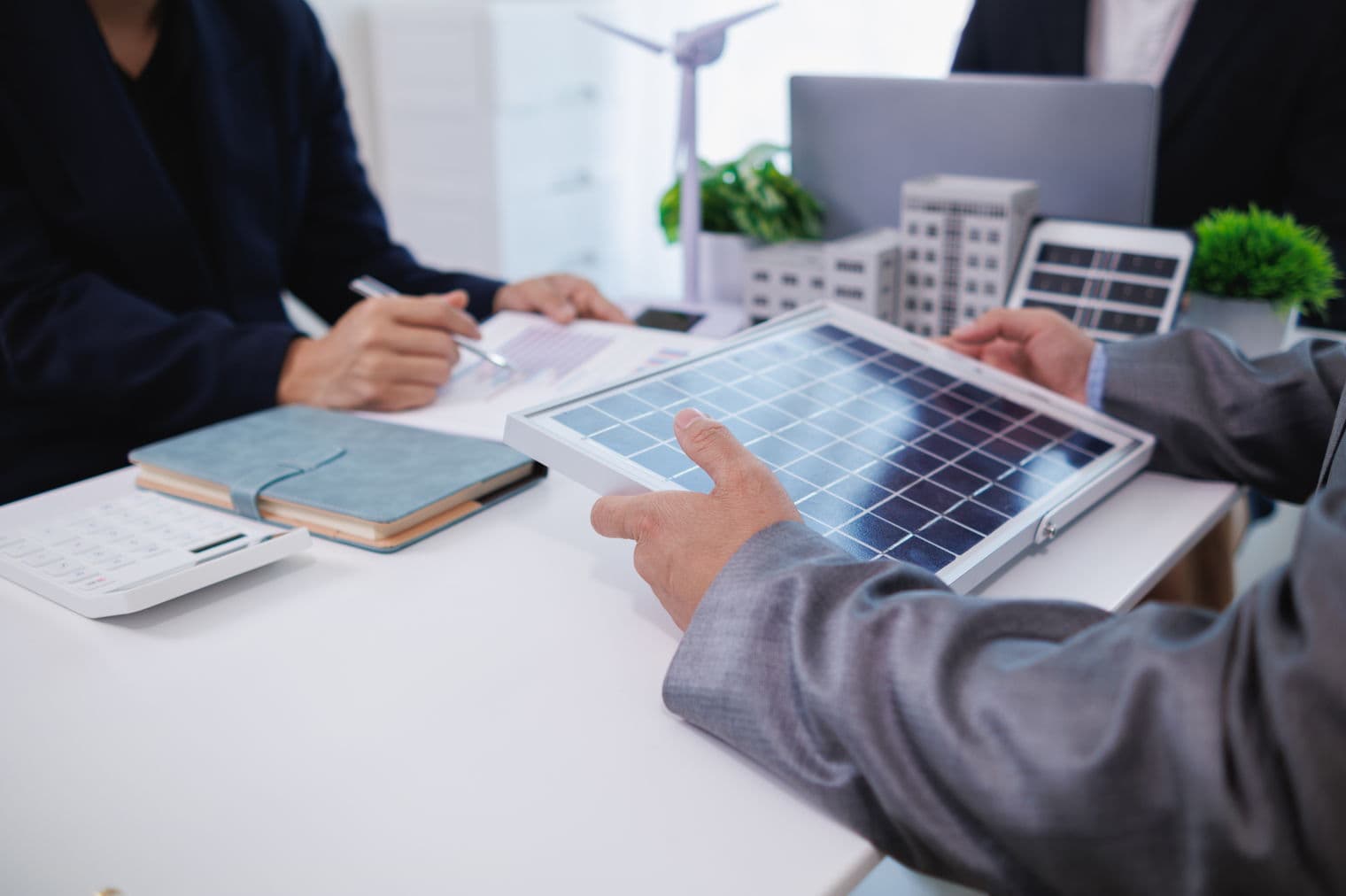 Zwei Personen in Anzügen diskutieren über ein Solarpanel an einem Tisch mit einem Taschenrechner, einem Notizbuch und Modellgebäuden.