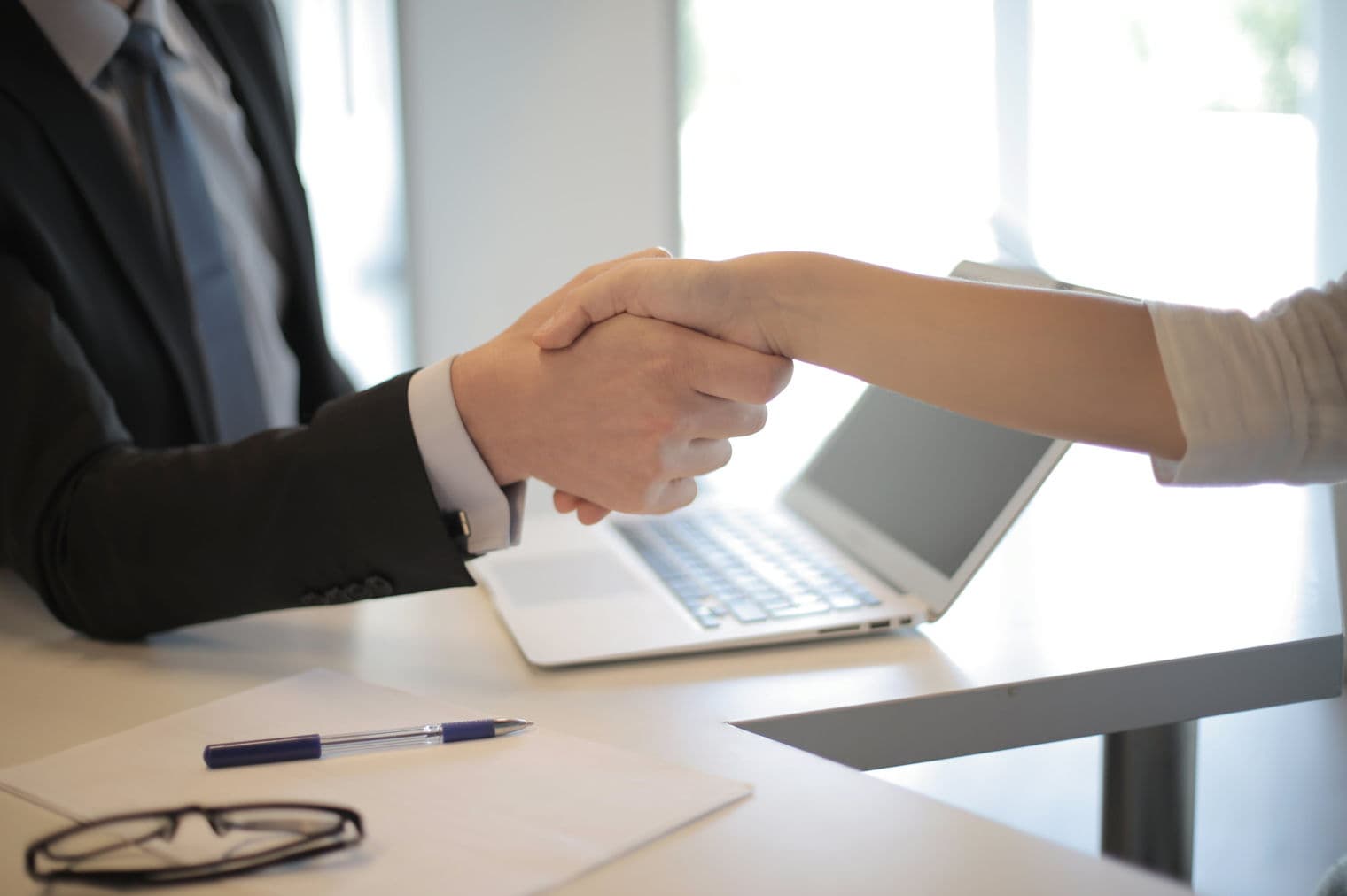 Two people shake hands over a white desk with a pen, paper, and glasses. One person wears a black suit and blue tie.