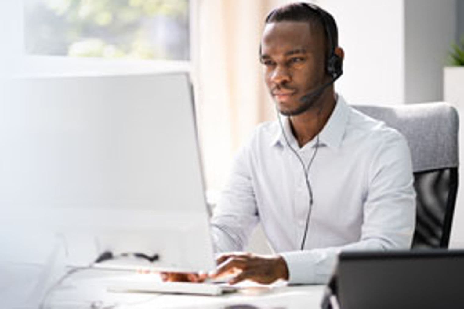 Mann mit Headset tippt an einem Computer in einem hellen Büro. Er trägt ein weißes Hemd und sitzt auf einem grauen Stuhl. Fenster im Hintergrund.