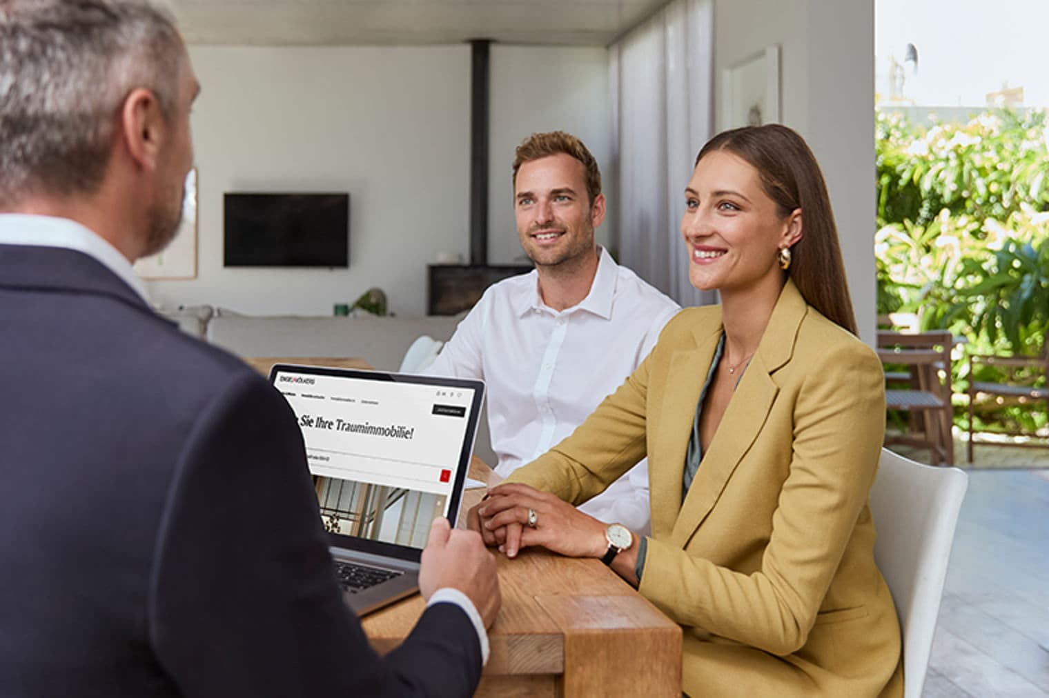 A real estate agent shows a couple properties on a laptop at a wooden table. The couple is smiling.