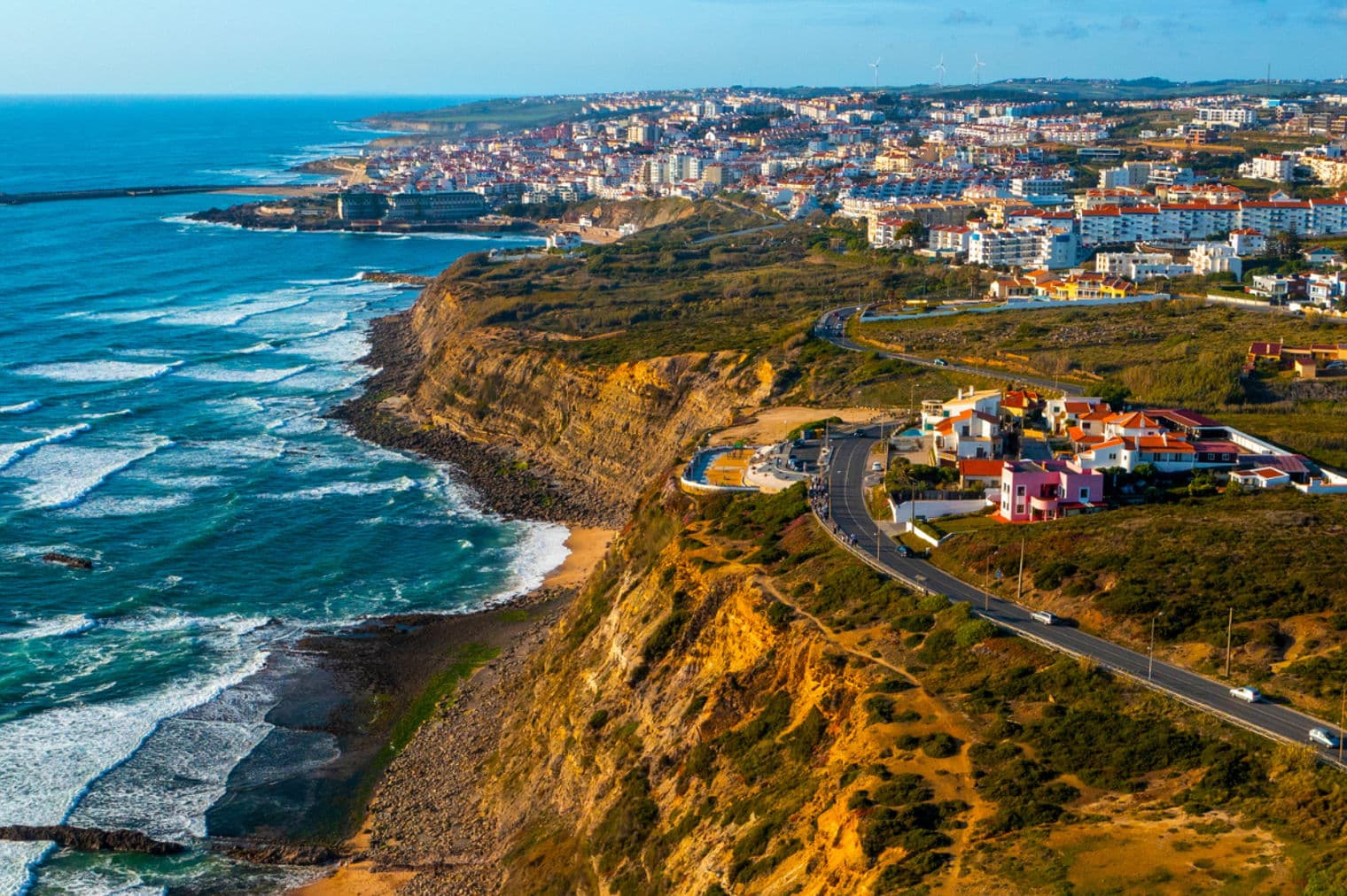 Luftaufnahme einer Küstenstadt mit weißen Gebäuden, einer gewundenen Straße entlang einer Klippe und blauen Meereswellen.