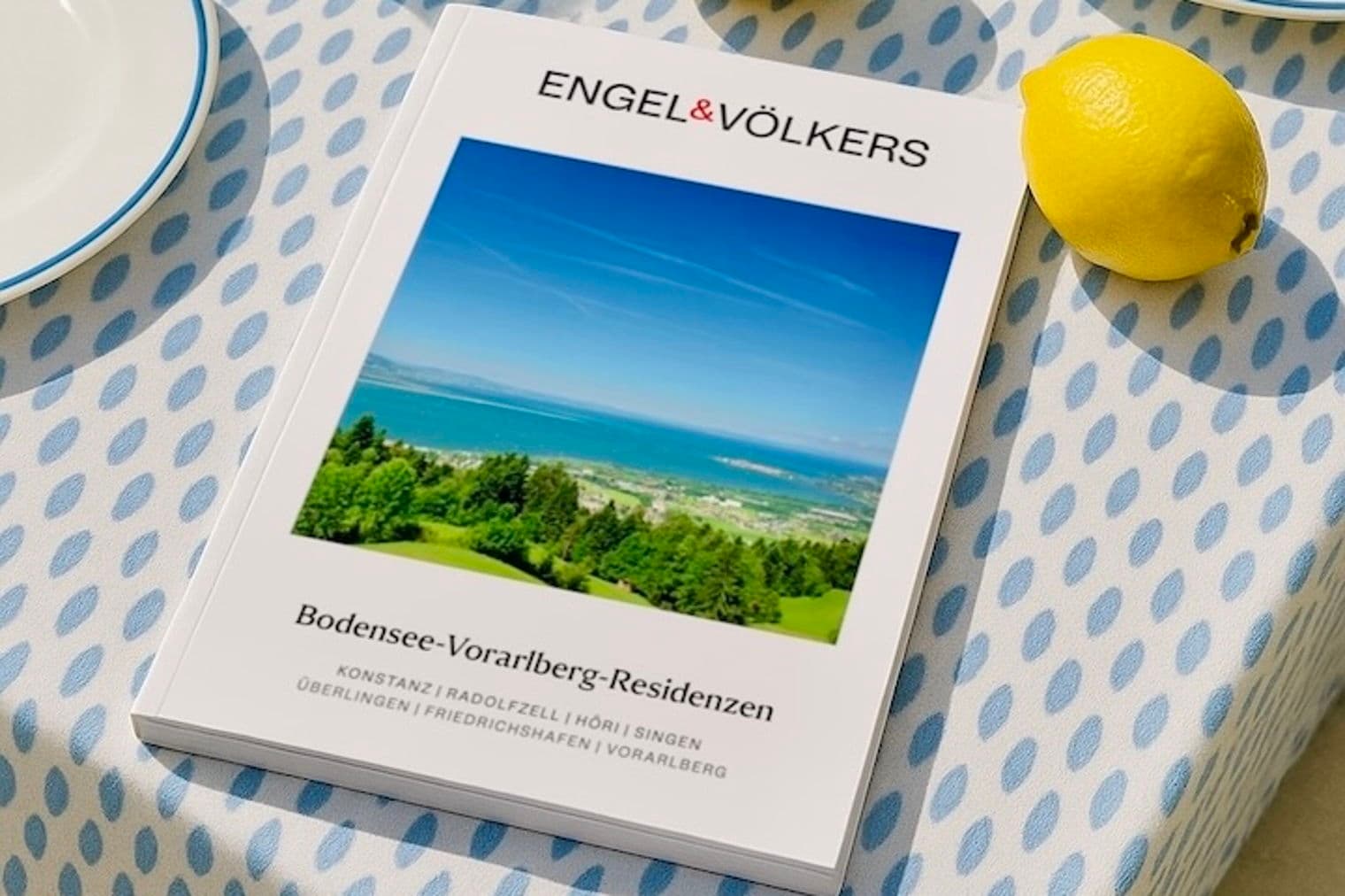 Engel & Völkers Immobilienkatalog auf einer blau gemusterten Tischdecke mit einer Zitrone und einem Teller.