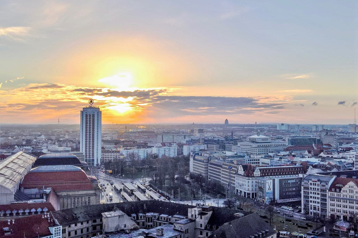 Stadtbild bei Sonnenuntergang. Ein hohes Gebäude mit einem "M"-Logo sticht vor dem orangefarbenen Himmel hervor. Gebäude und Straßen füllen den Vordergrund.