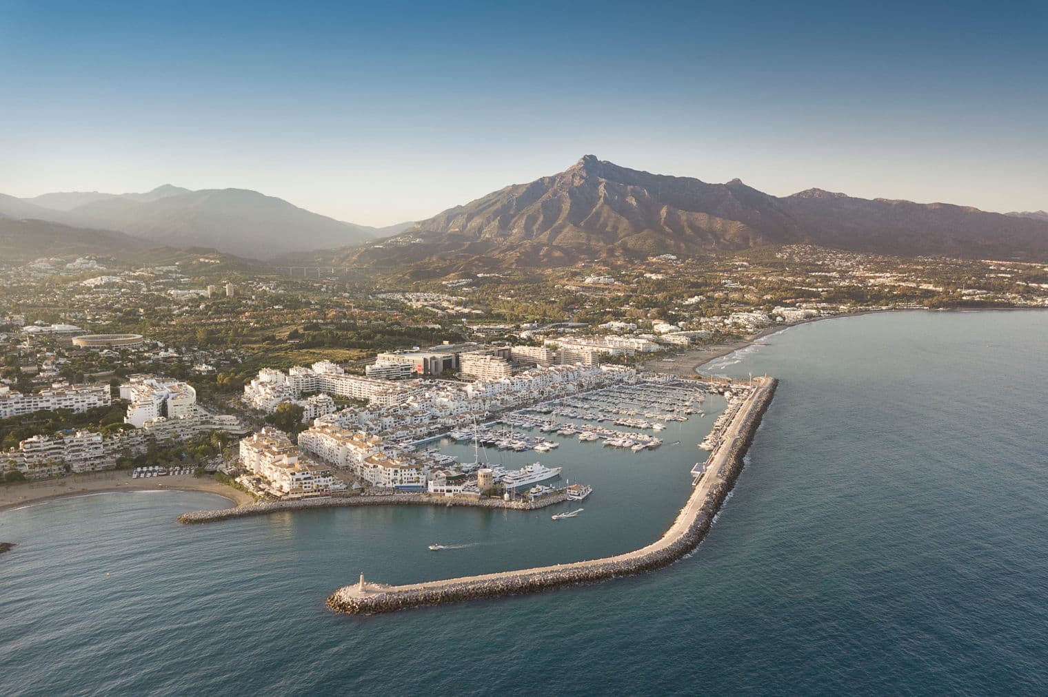 Vista aèria d'un port esportiu ple de vaixells, envoltat d'edificis blancs i muntanyes sota un cel blau.