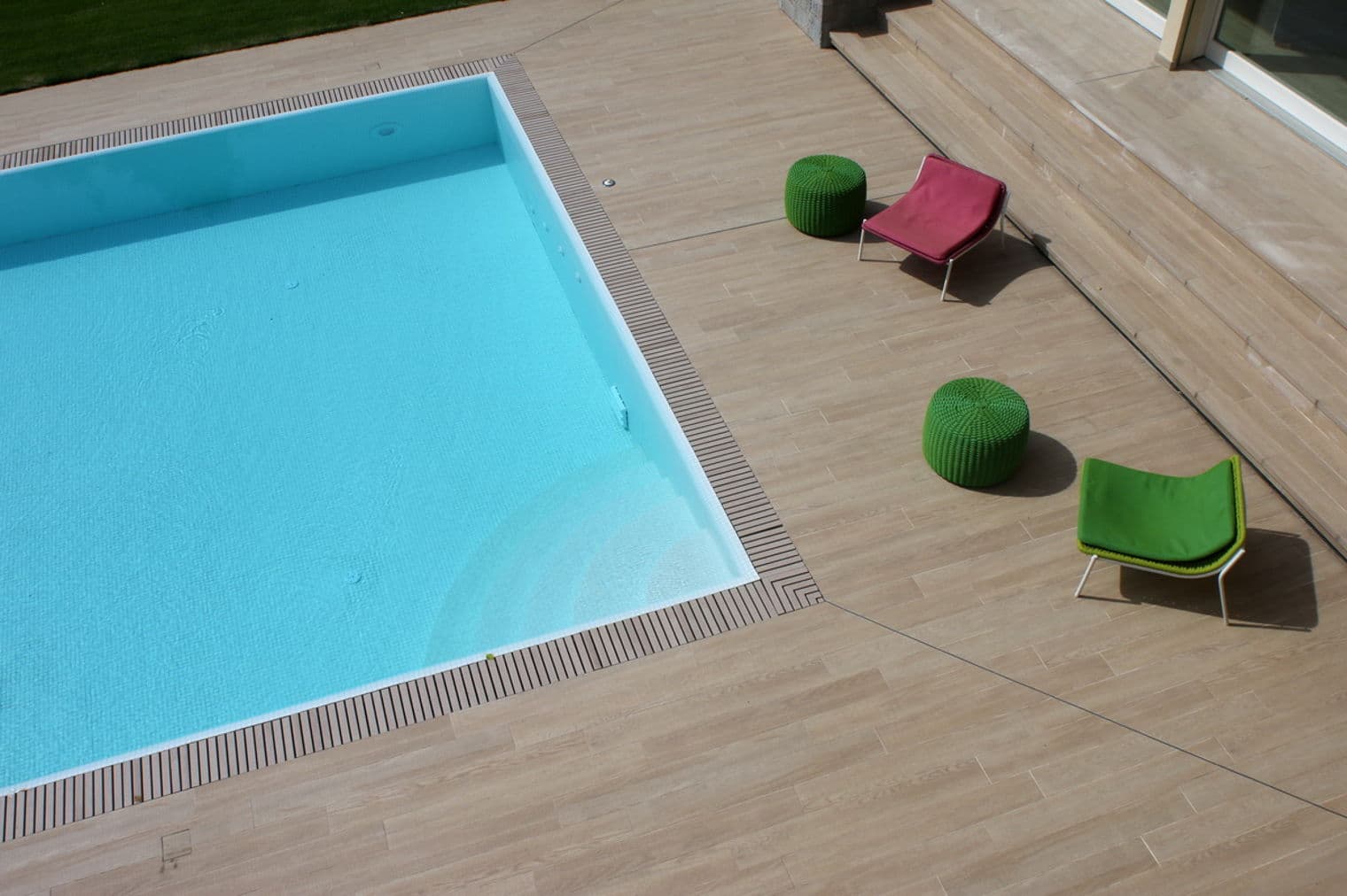 Aerial view of a pool with turquoise water, surrounded by a wooden deck with colorful chairs and poufs.