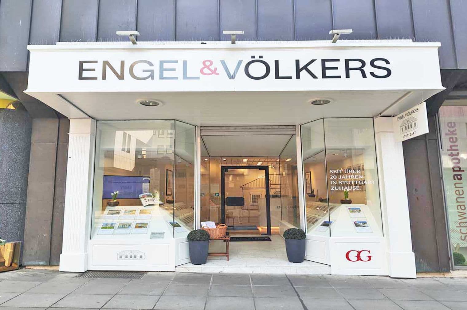 Engel & Völkers real estate storefront with glass windows displaying property listings. A basket sits on a small table near the entrance.