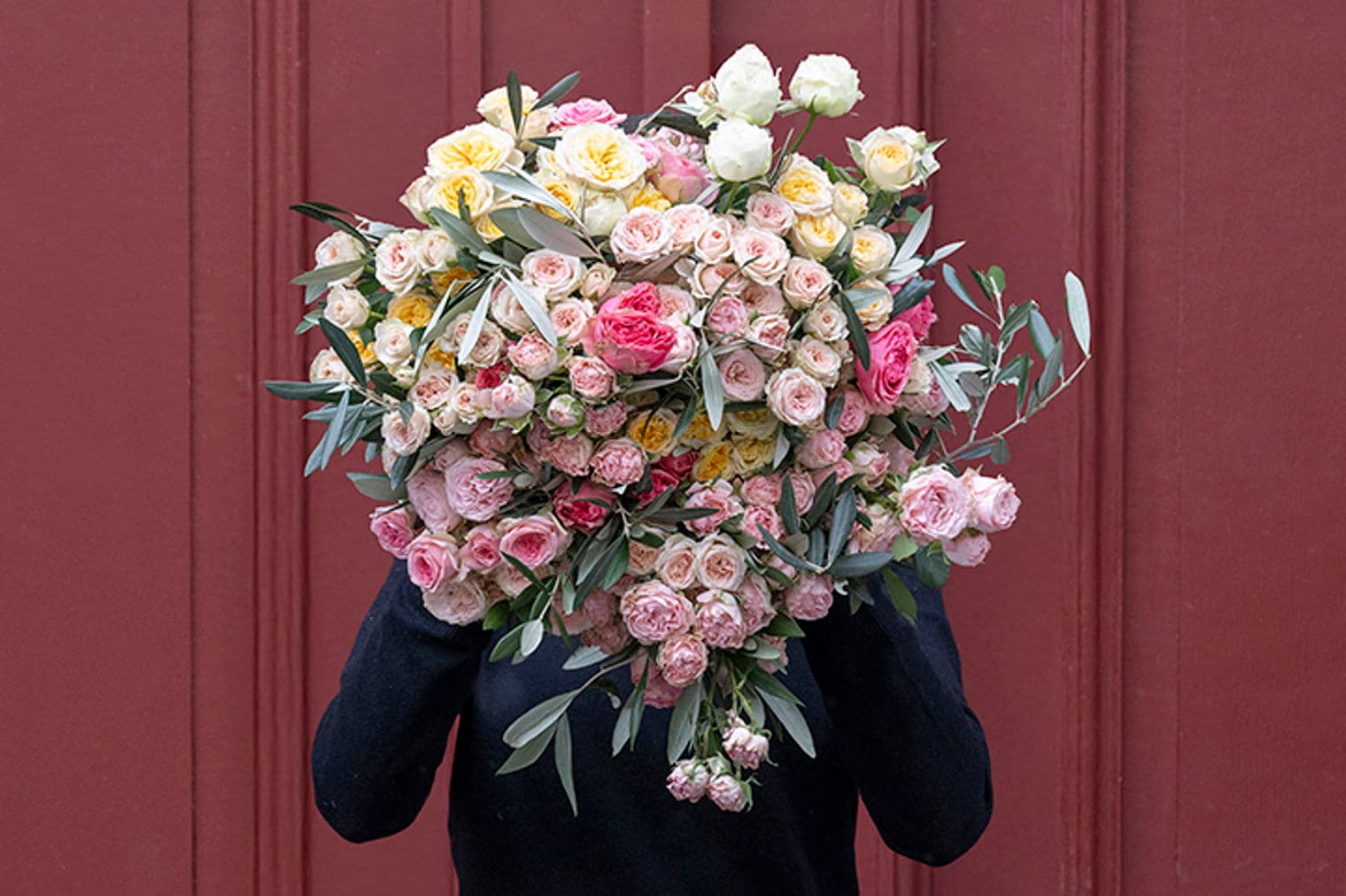 Person in schwarzem Pullover hält einen großen Strauß rosa, gelber und weißer Rosen vor einer roten Tür.