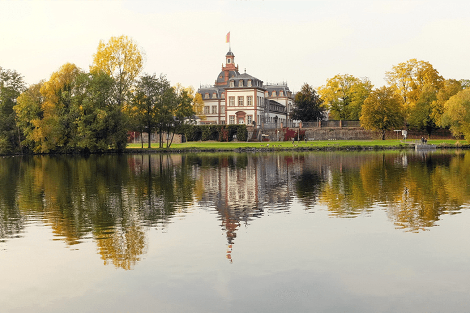 Malerische Aussicht auf ein weißes und rotes Herrenhaus mit einer Flagge, das sich in einem ruhigen See spiegelt, umgeben von Herbstbäumen.
