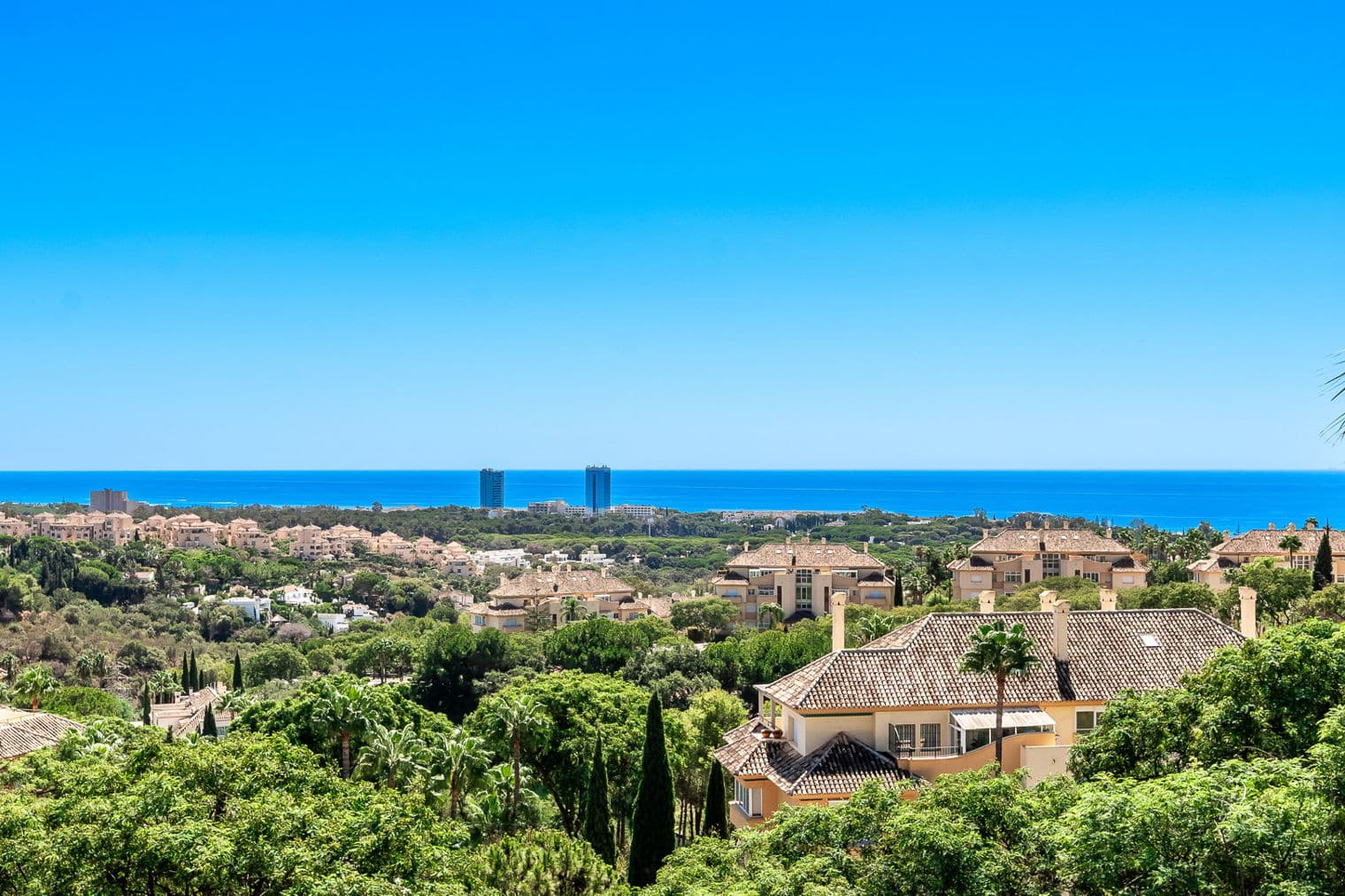 View of a coastal community with terracotta-roofed buildings, green trees, and the blue ocean under a clear sky.