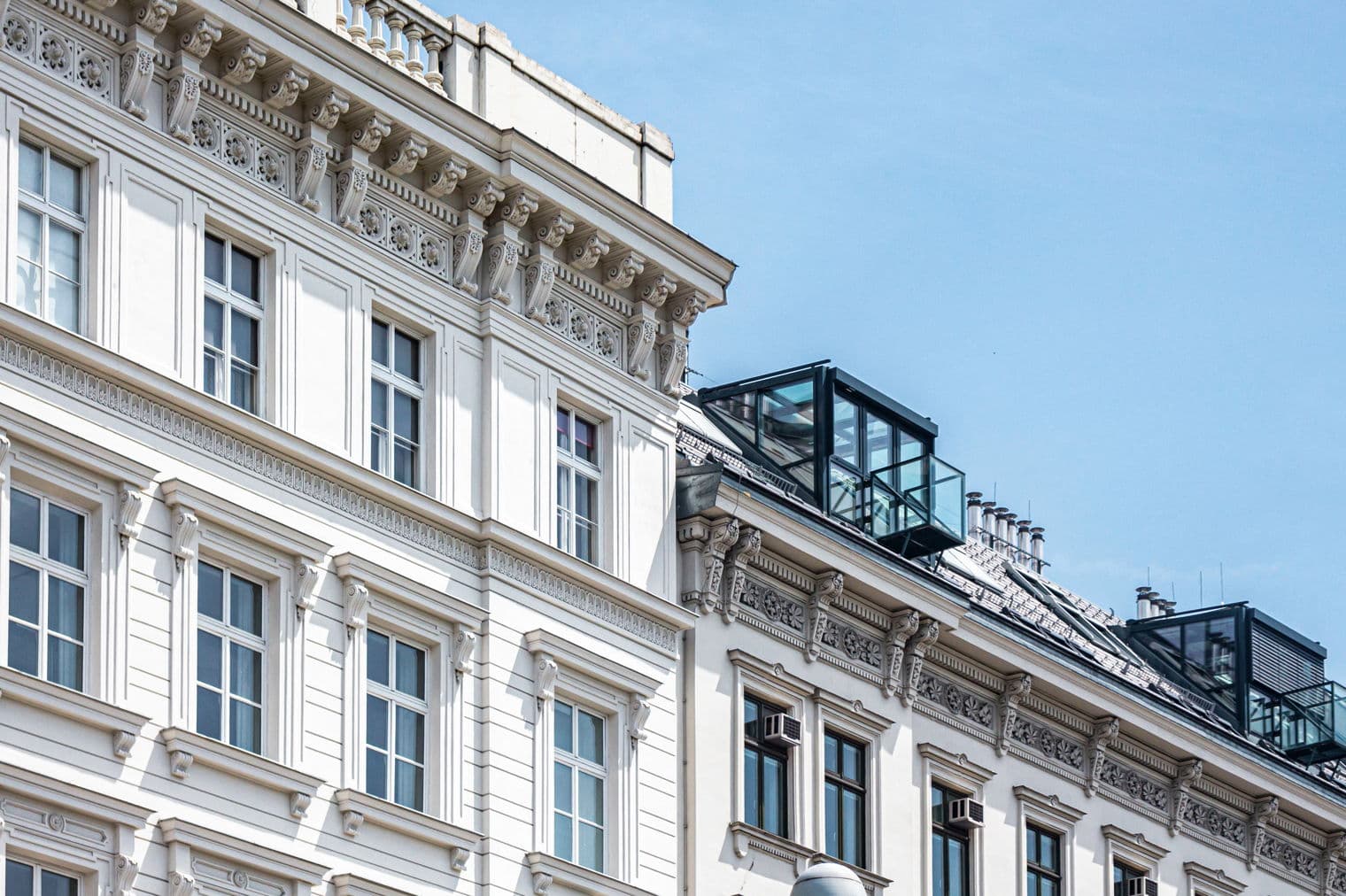 Exterior view of two ornate, white buildings against a blue sky. The buildings have decorative trim and dark-framed windows.