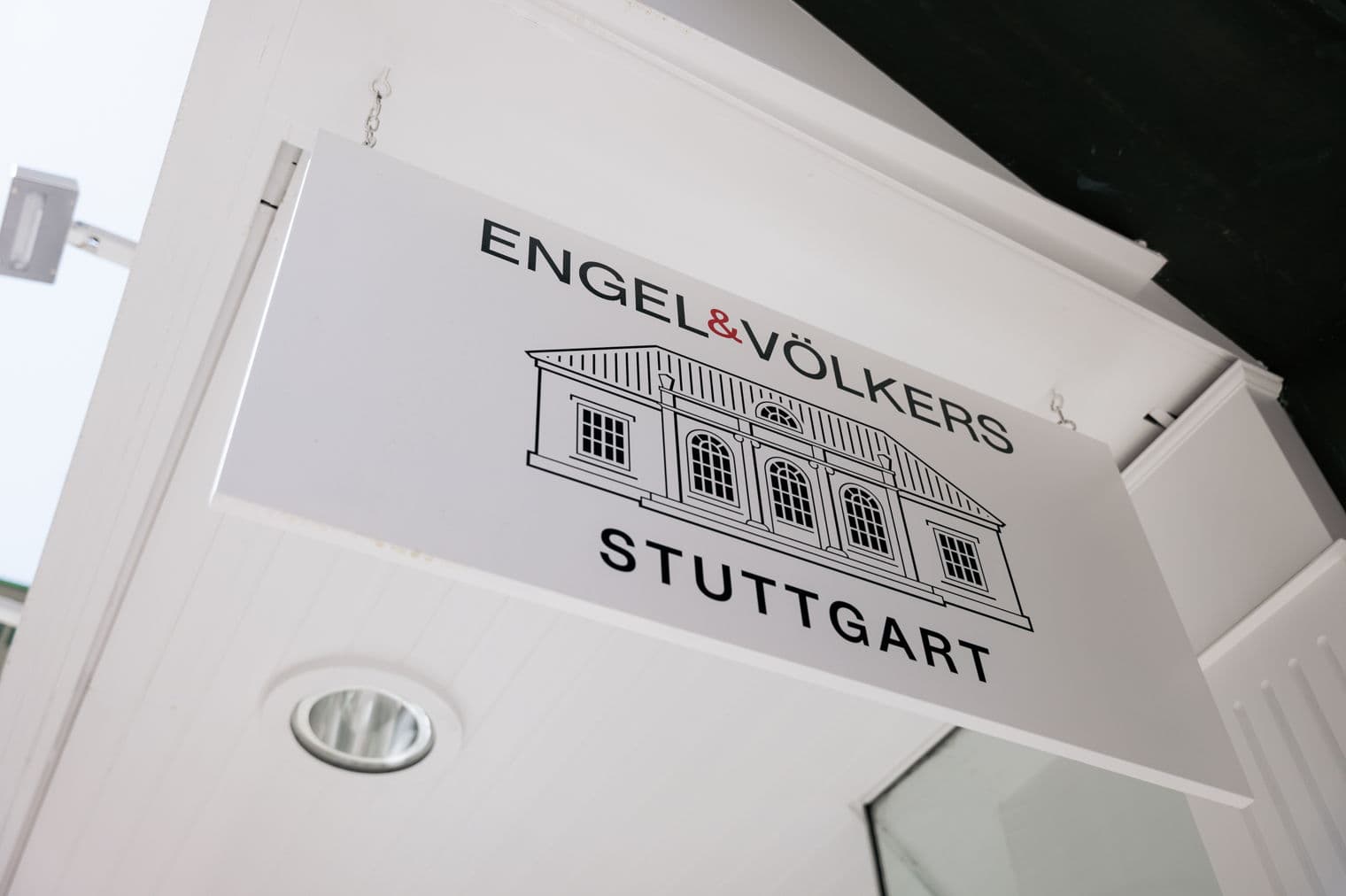 A white Engel & Völkers sign hangs from a white building. The sign reads "Stuttgart" below a building outline.